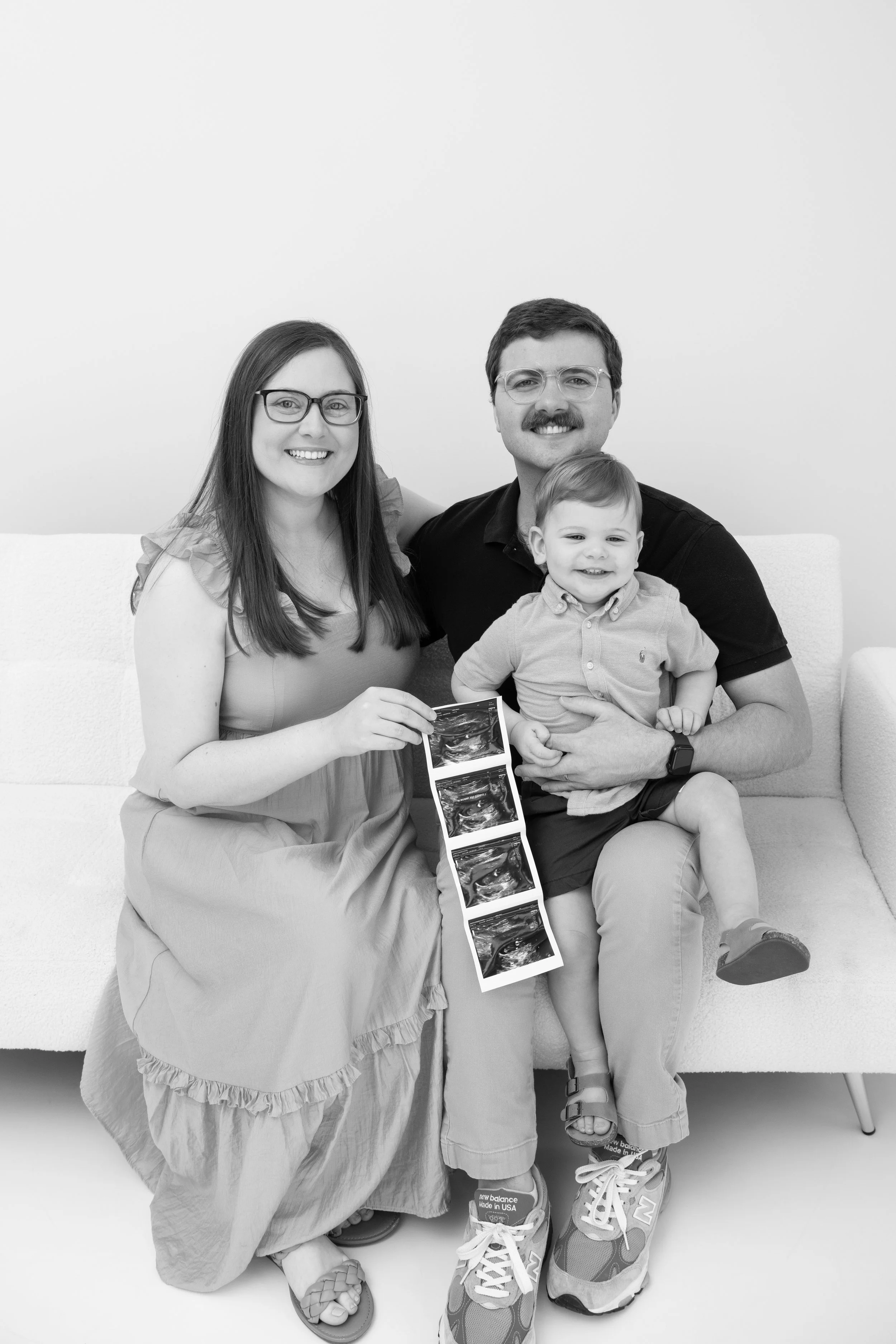 Family of three with a woman holding ultrasound images, sitting on a couch, with a man holding a young boy. All smiling at the camera.