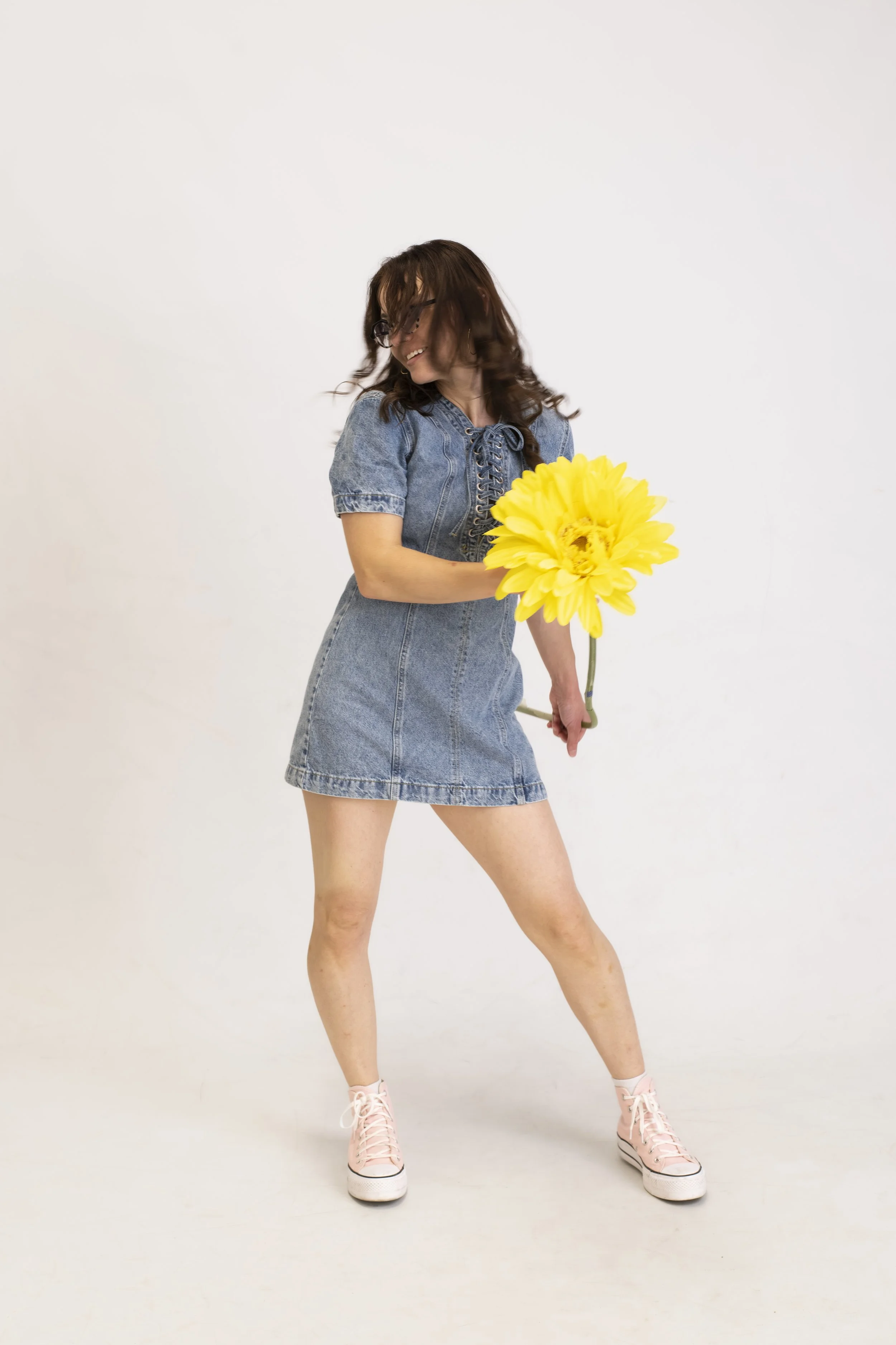 Woman in a denim dress holding a large yellow flower while smiling and looking to her left, standing against a plain white background.