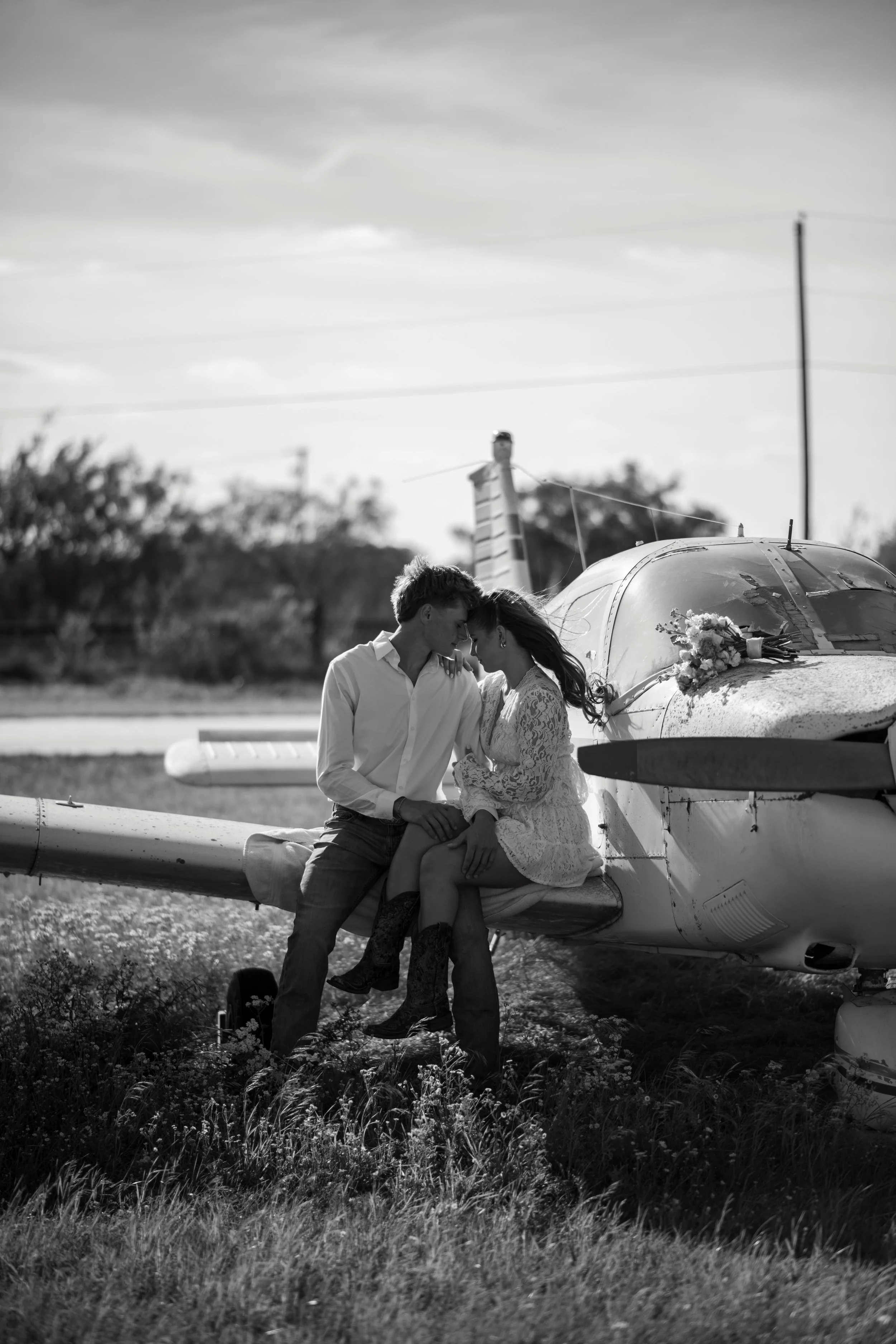 Black and white photo of a couple sitting on the wing of a small airplane in an open field, with the woman wearing a lace dress and the man in a white shirt. The woman has flowers in her hair, and the couple is leaning their heads together, holding h