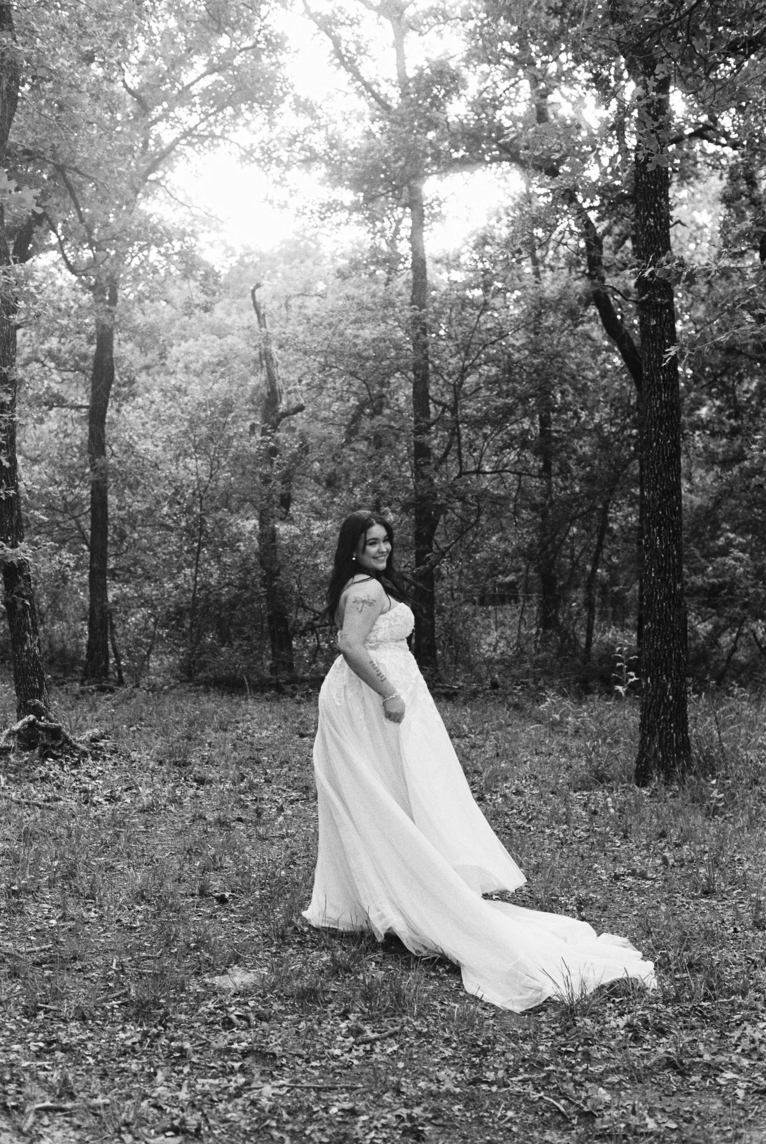 A woman wearing a wedding dress standing in a wooded area with trees and sunlight in the background.