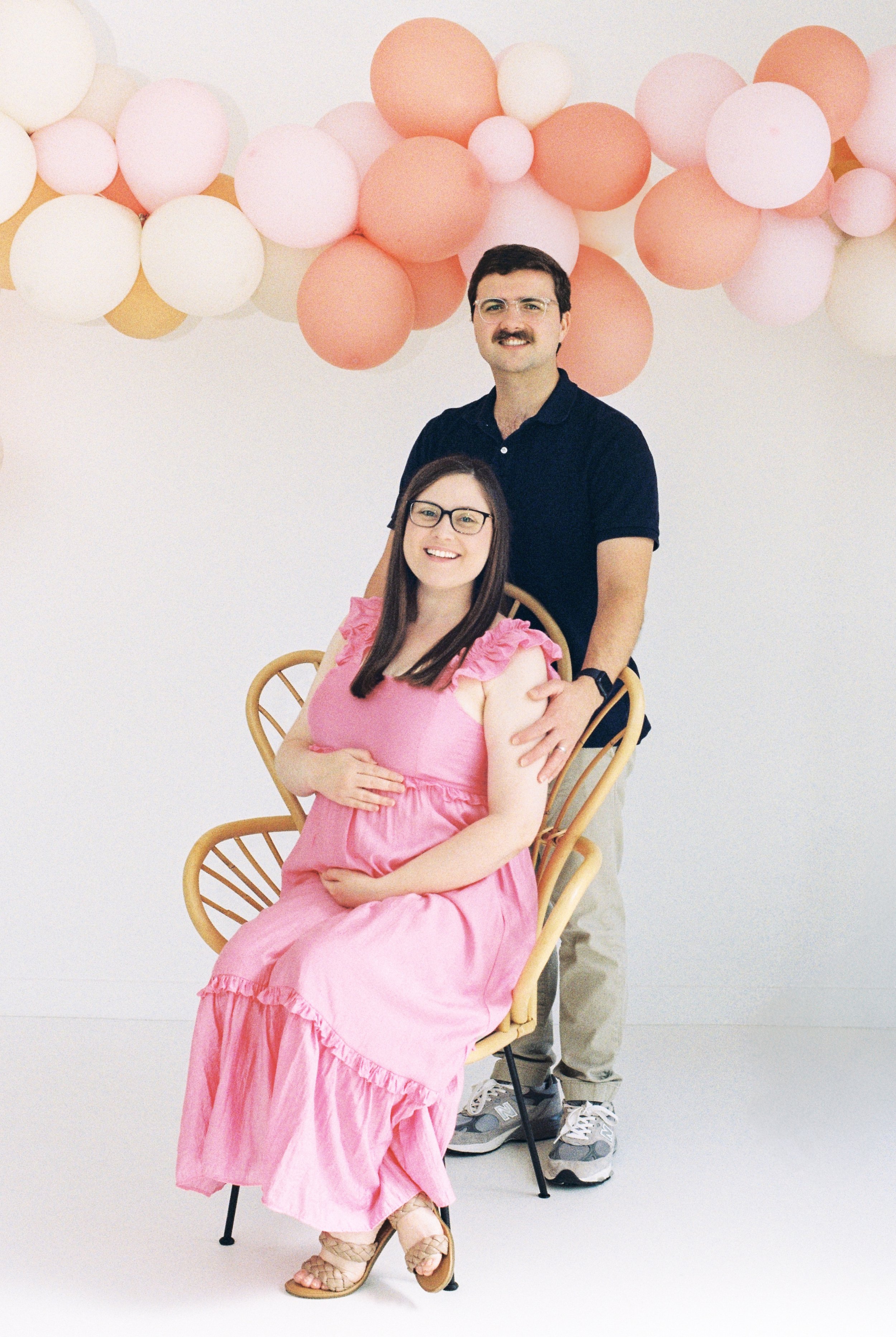 A woman in a pink dress sitting in a yellow chair, smiling, with a man standing behind her, both posing in front of a background with pink and white balloons.