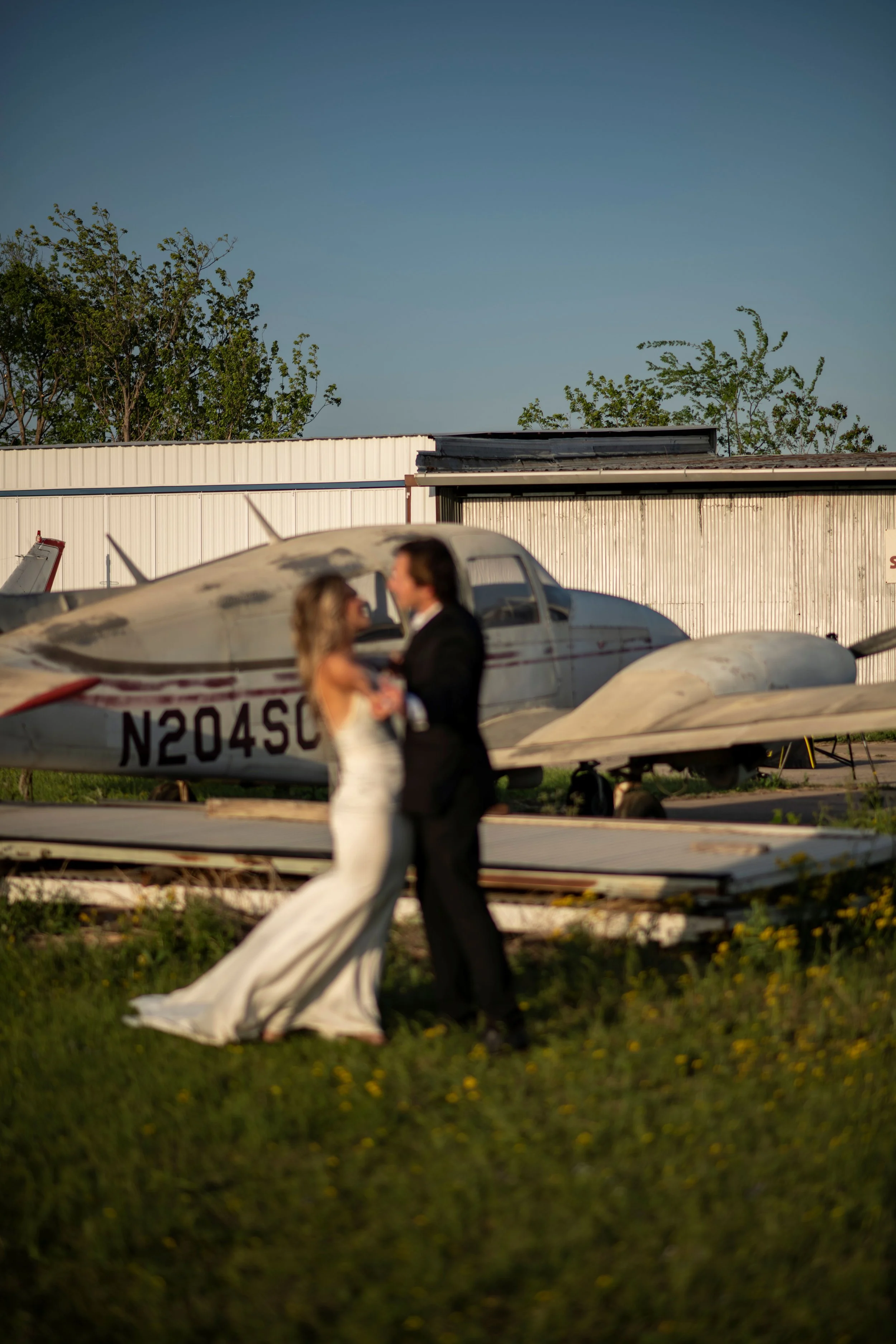 A blurred couple dressed in formal attire walking near a small aircraft in an outdoor area at dusk, with trees and a metal building in the background.