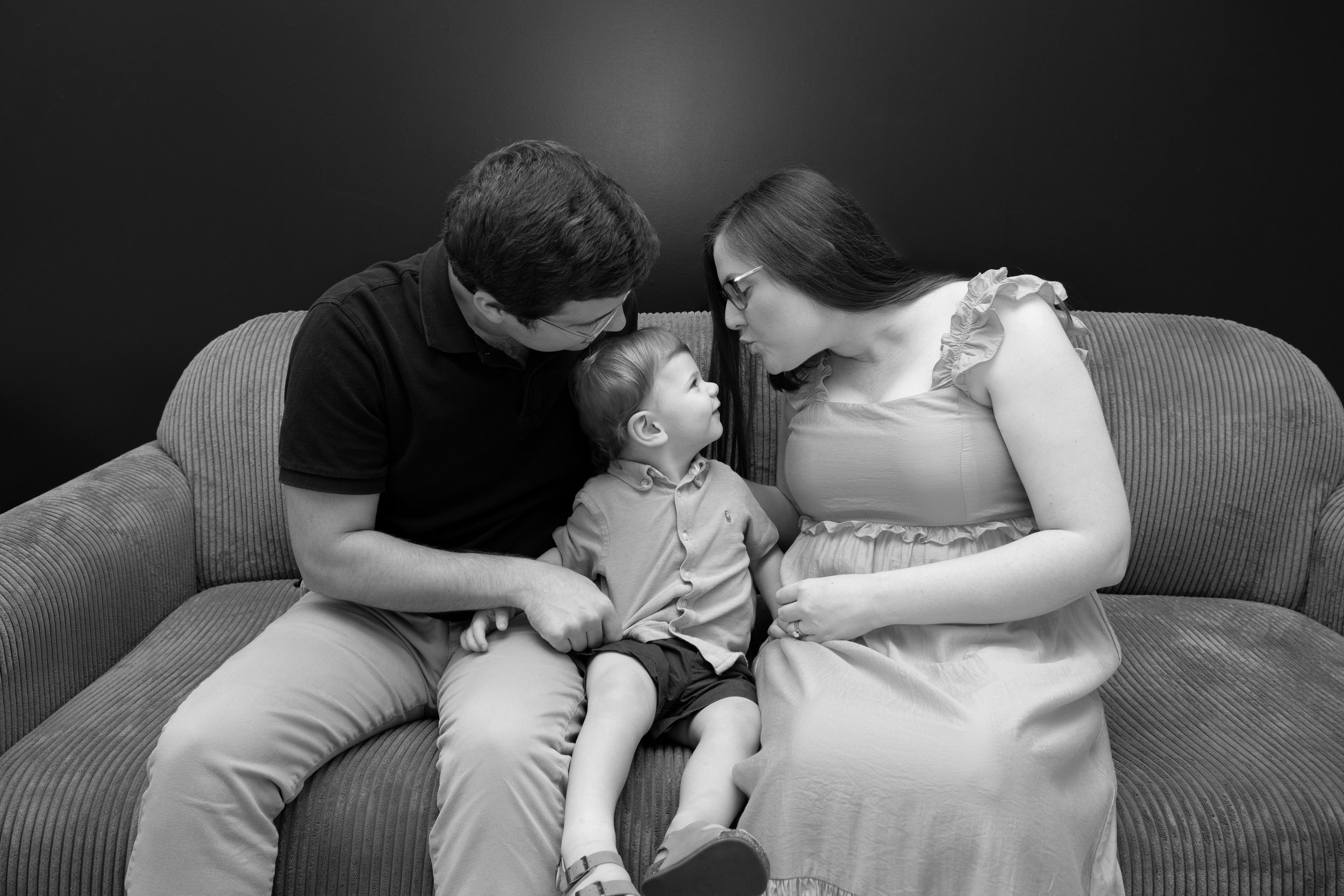 A family of three sits on a sofa, with a father, mother, and young son, all leaning in close and making eye contact.