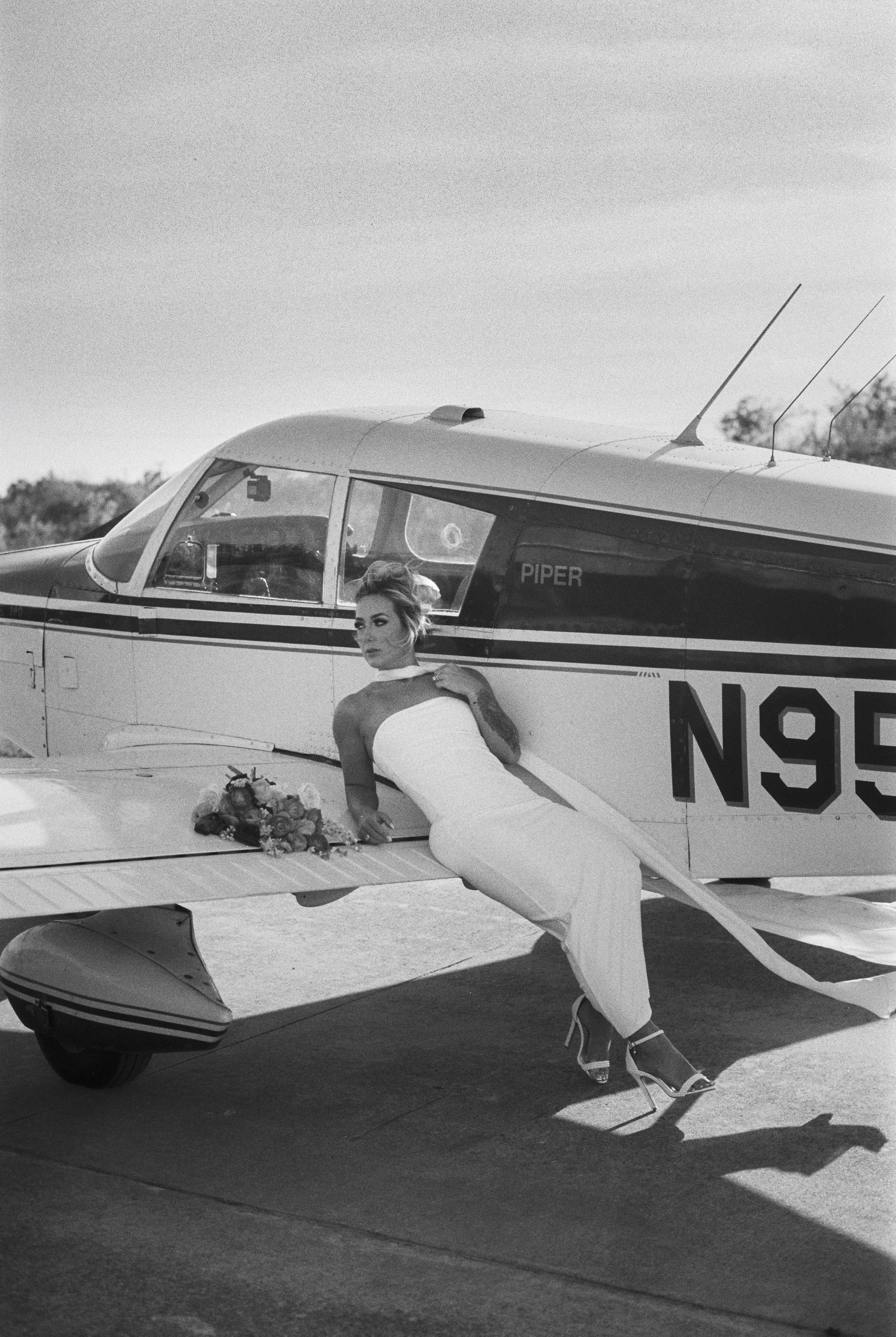 Black and white photo of a woman standing beside a small airplane with the name 'Piper' on it, holding a bouquet of flowers, with her right hand resting on the airplane and her left foot on the plane's landing gear.