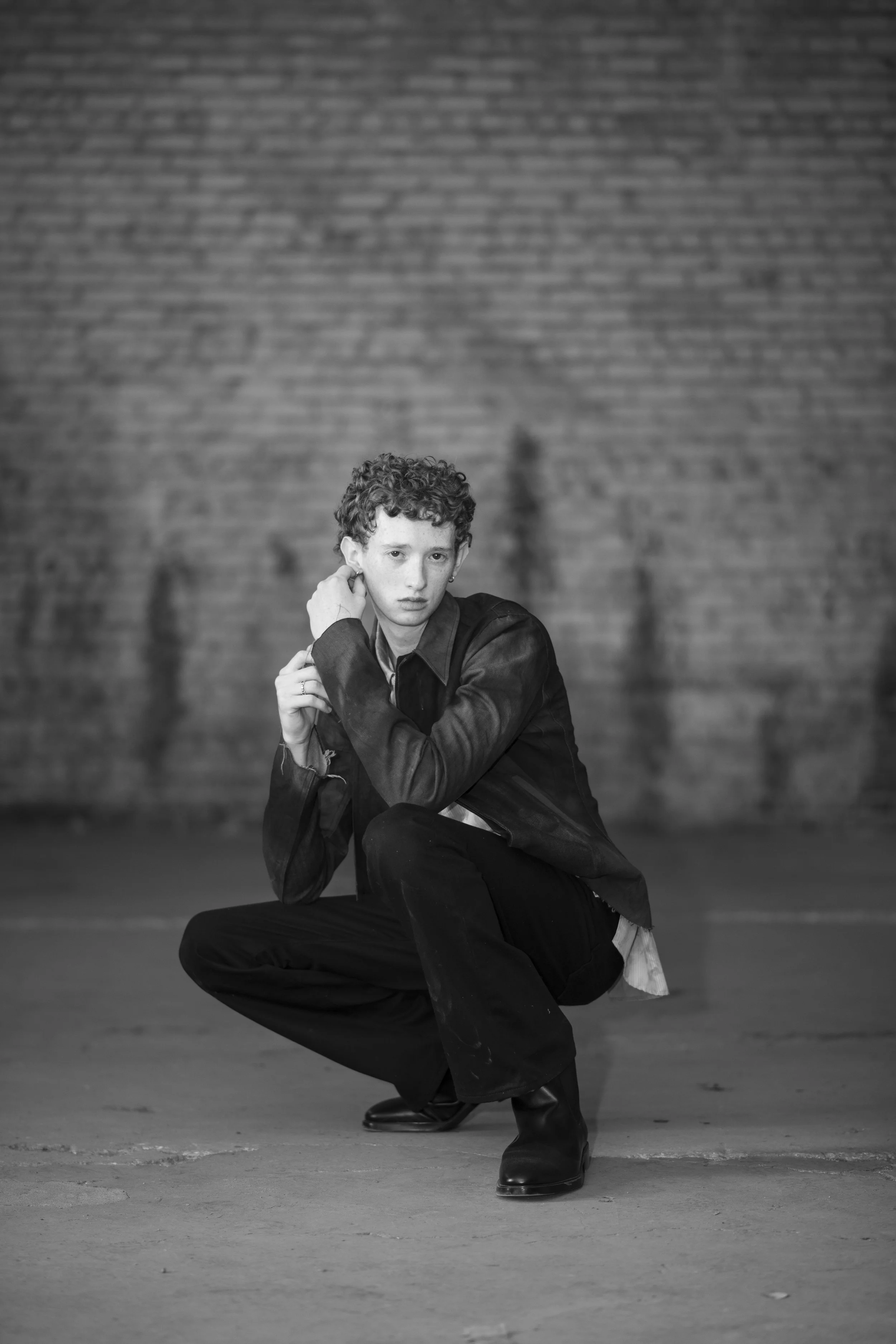 Black and white photo of a young person with curly hair, crouching down in front of a brick wall, wearing a dark jacket and boots, with a serious expression.