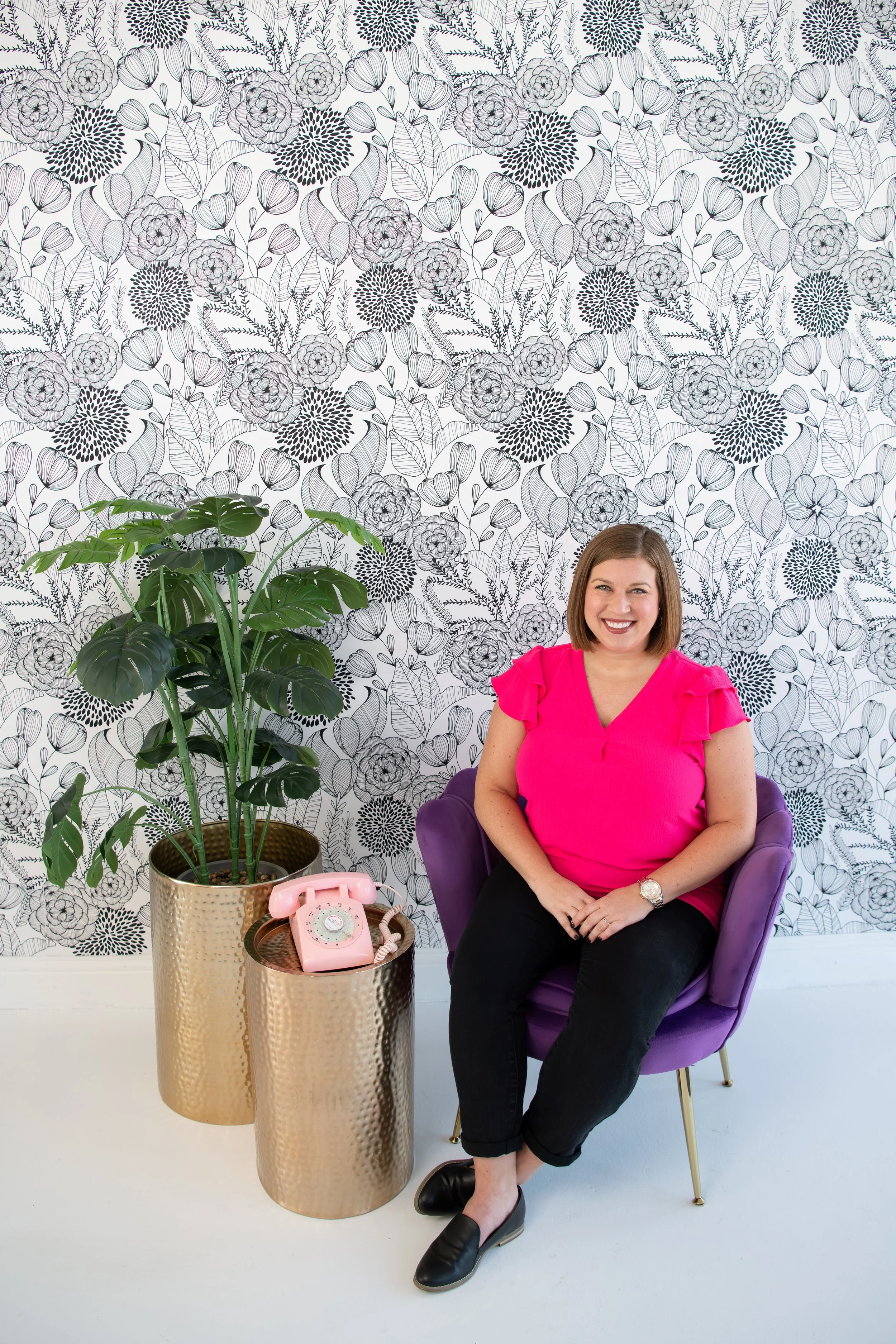A woman with shoulder-length brown hair, sitting in a purple armchair, smiling and wearing a bright pink top, black pants, and black shoes. She is in a room with a floral black-and-white patterned wallpaper in the background, a large green potted pla