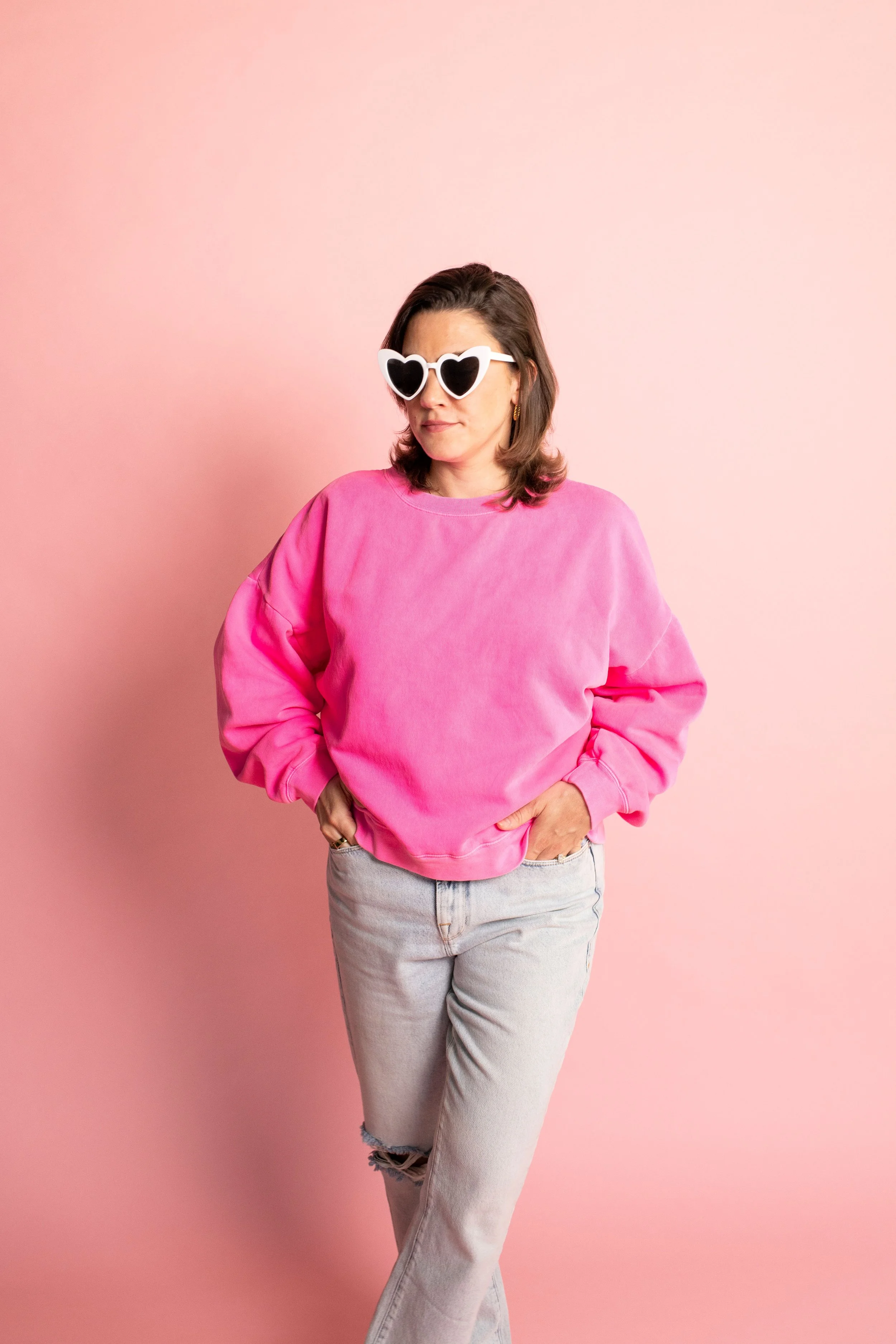 Woman in pink sweatshirt and ripped jeans wearing heart-shaped sunglasses poses against pink background.