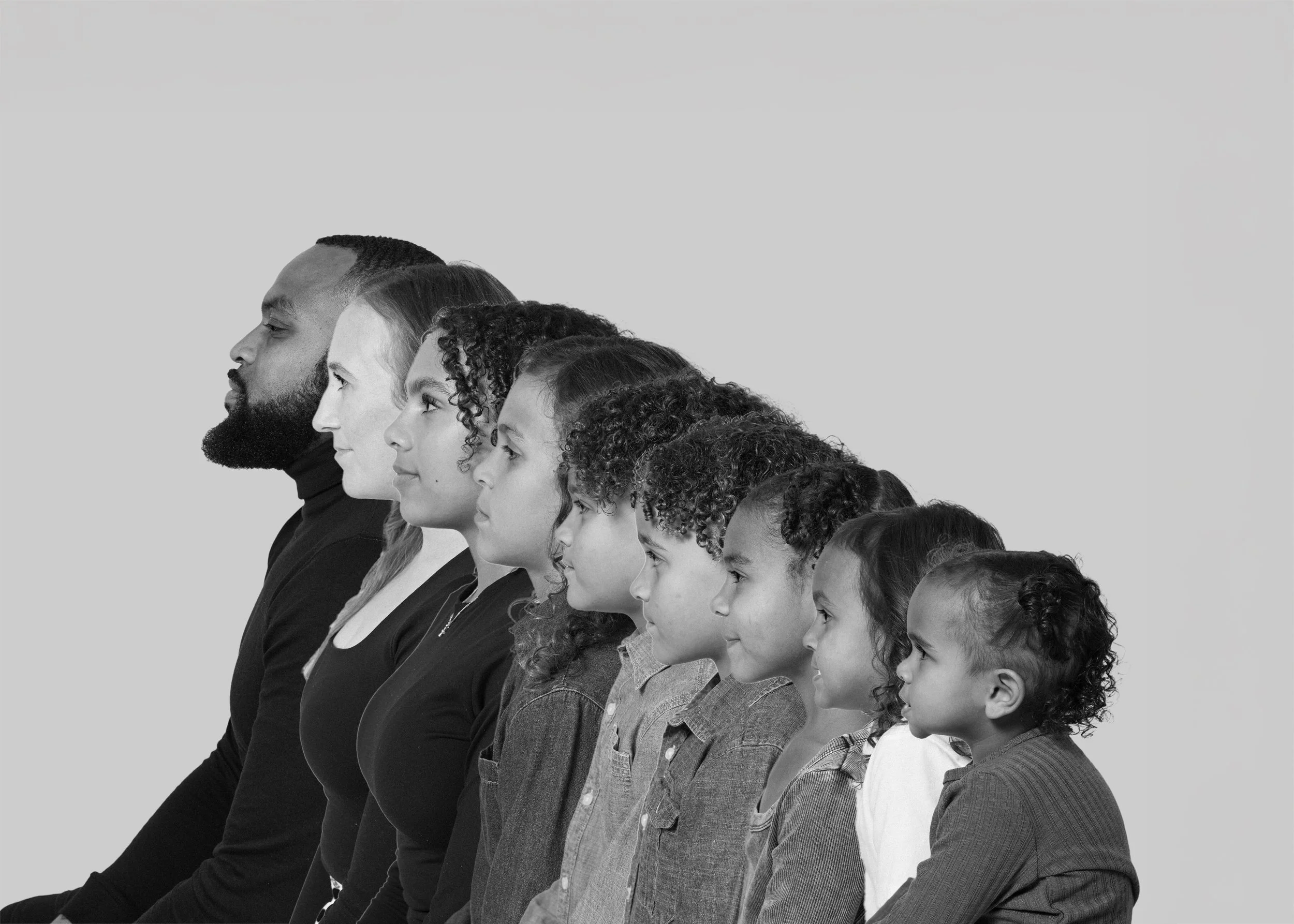 A black and white photo of a diverse group of children and adults sitting in a line, facing left with calm expressions.