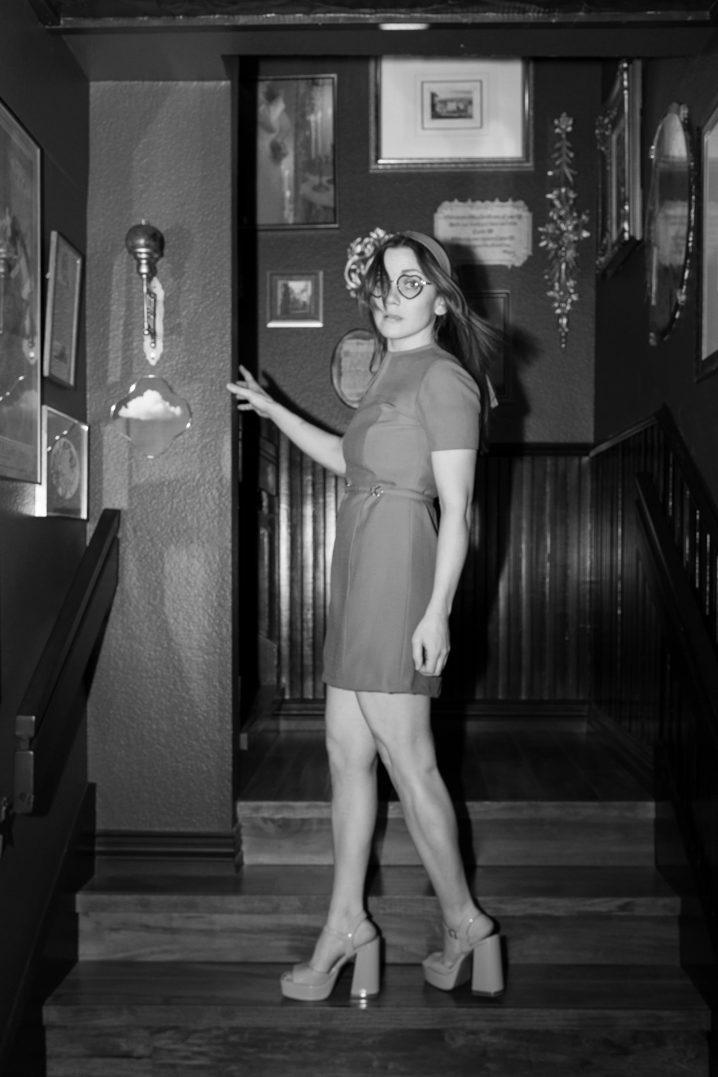 A woman with glasses and shoulder-length hair standing on a staircase, wearing a short dress and high-heeled shoes, in a home decorated with framed pictures, artwork, and hanging ornaments.