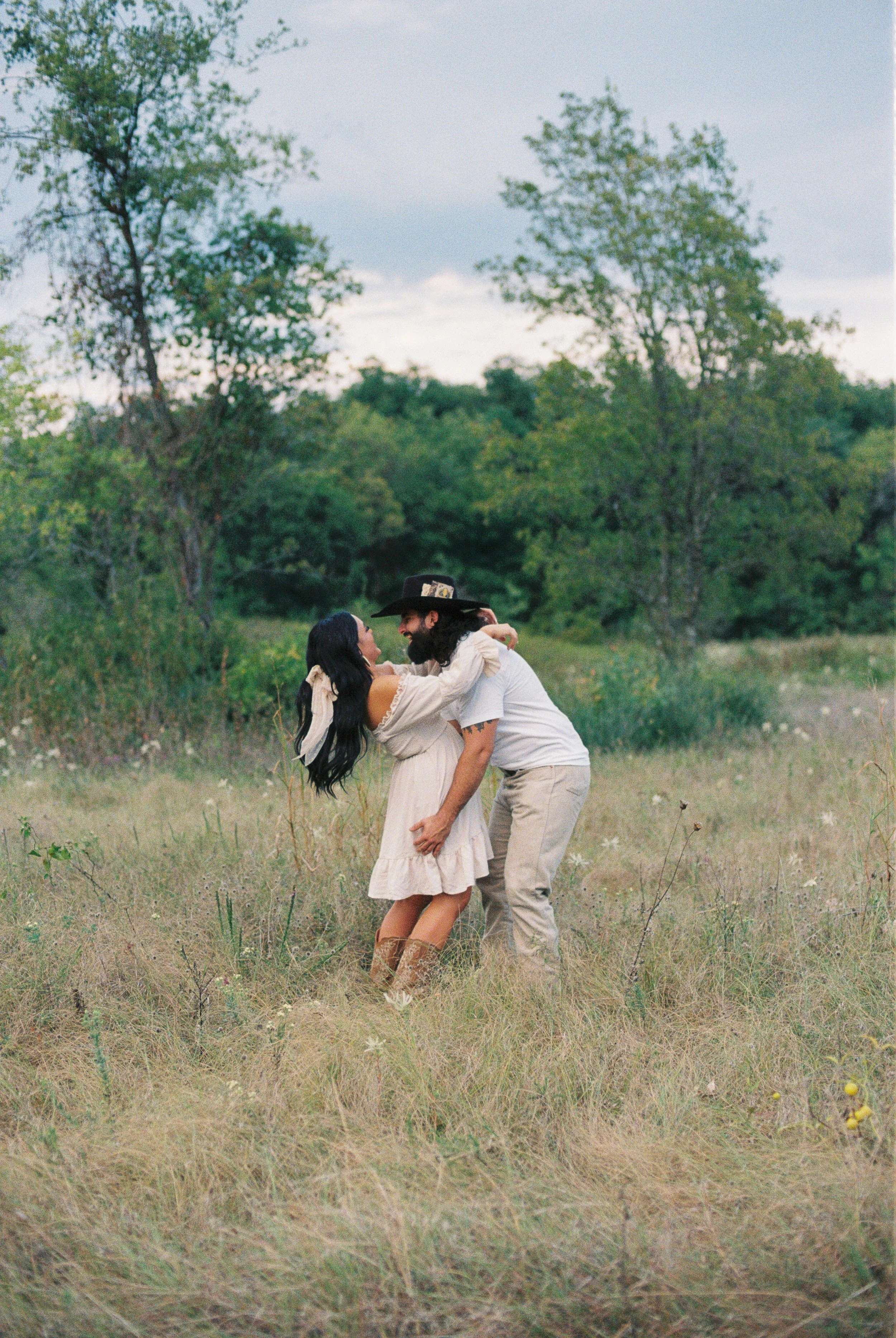 A couple dancing and embracing in a grassy field with trees in the background, one wearing a black cowboy hat and the other a cream-colored dress.