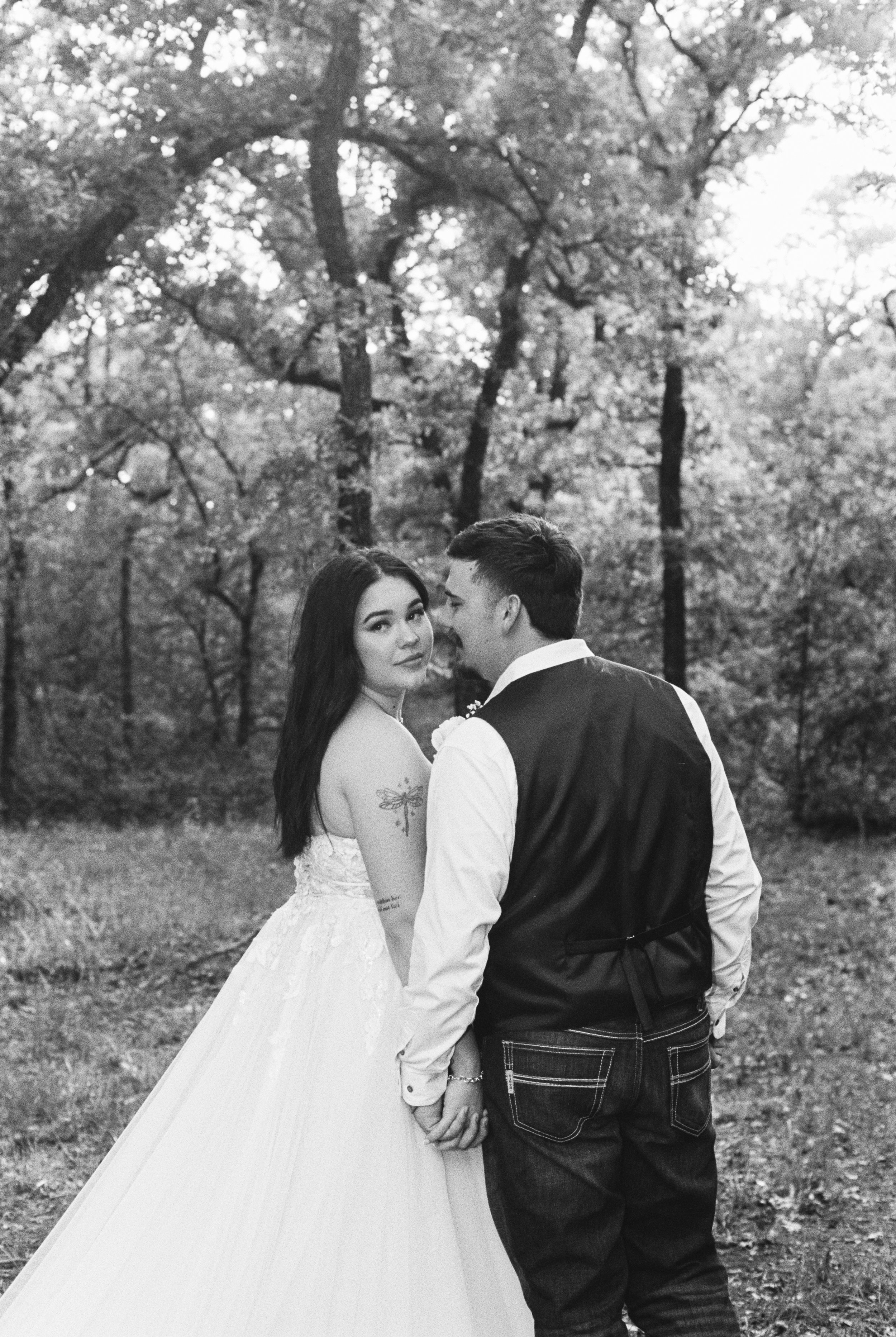 A black and white photo of a couple in a park surrounded by trees. The woman is in a wedding dress and the man is in a vest and jeans. They are smiling and holding hands.