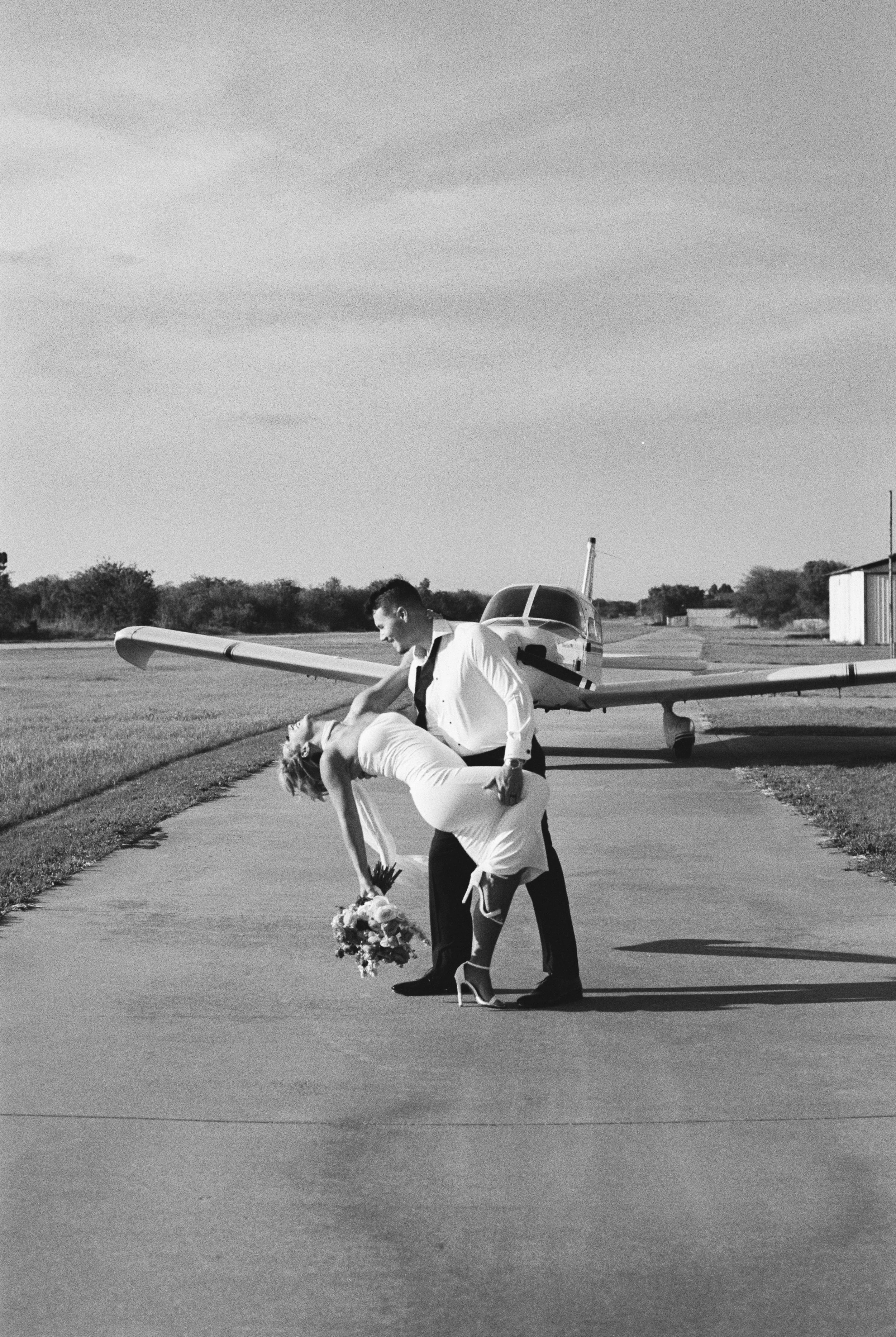 A bride and groom kissing during their wedding ceremony outdoors on a paved path, with an airplane in the background.