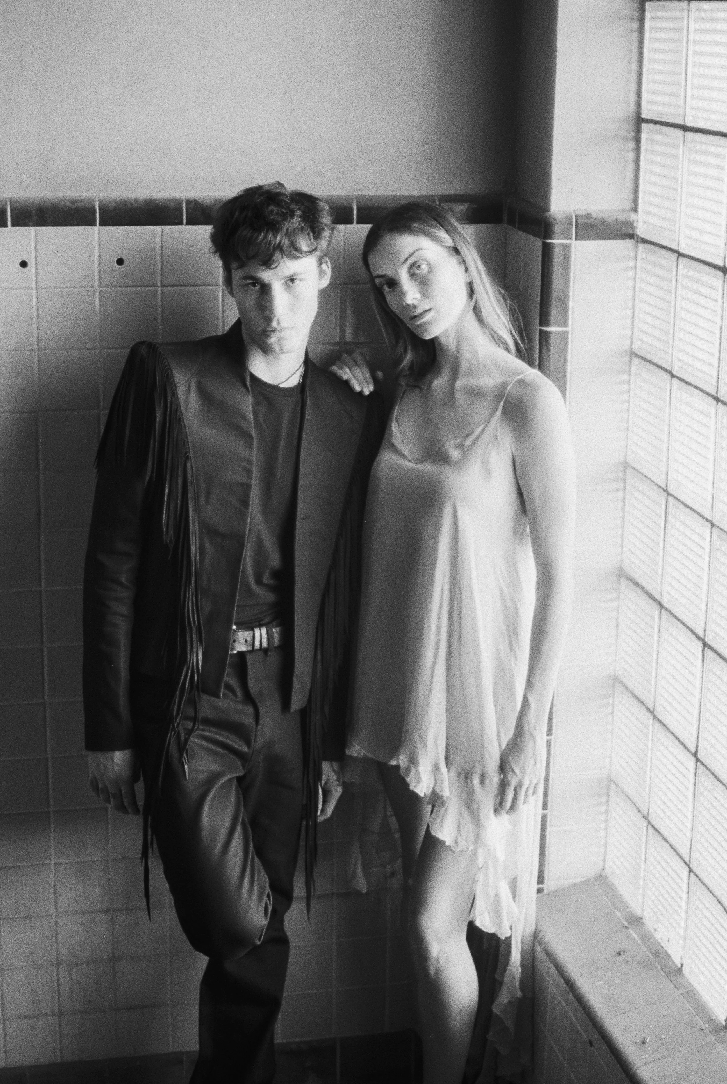 Young man with wavy hair dressed in a leather jacket and dark pants, standing with arms crossed, next to a life-size skeleton wearing a dress, in a tiled room with a corner of the room visible.