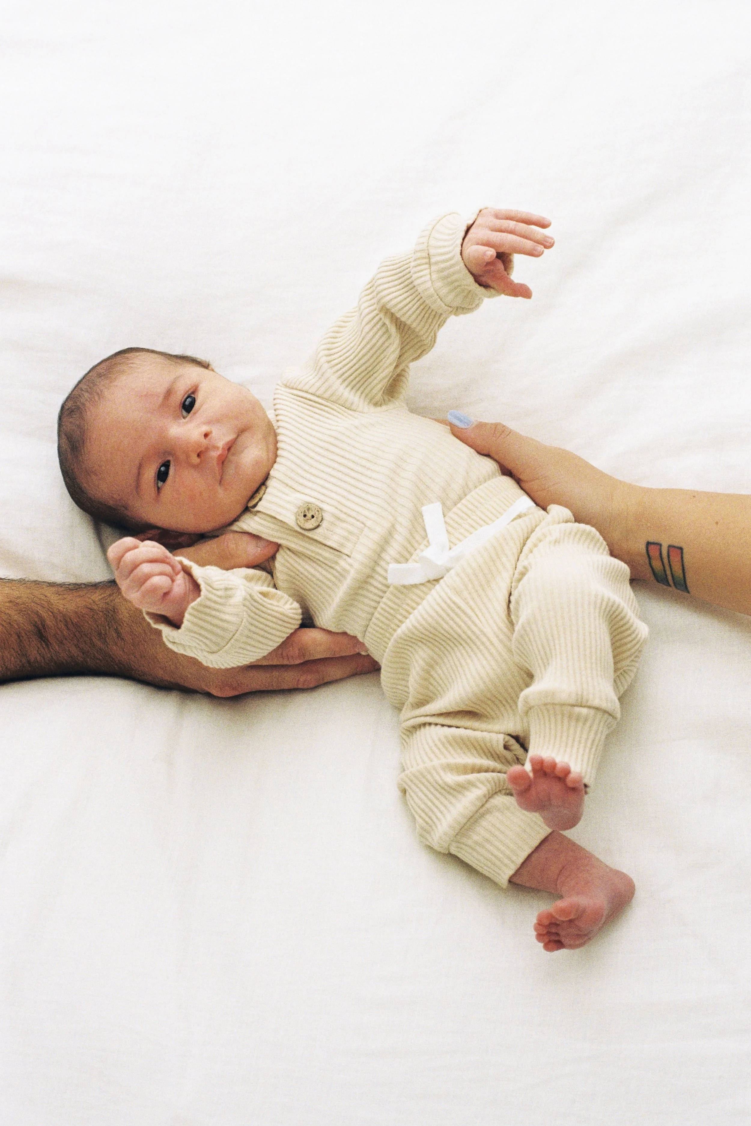 A baby lying on a white bed with support from an adult's hand, looking at the camera, wearing a cream-colored ribbed outfit and surrounded by soft lighting.