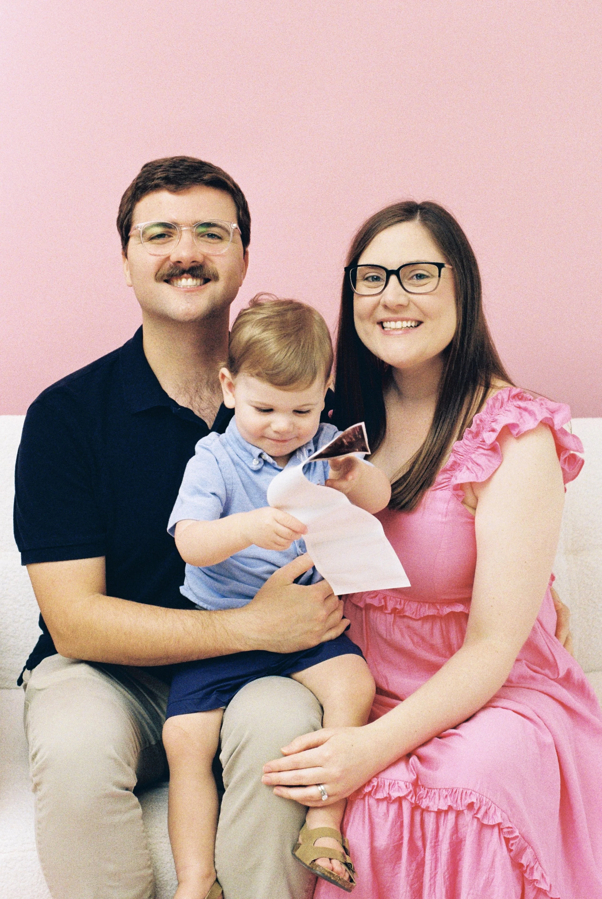 A smiling family of three sitting on a beige couch in front of a pink wall, looking at a photograph or piece of paper together.