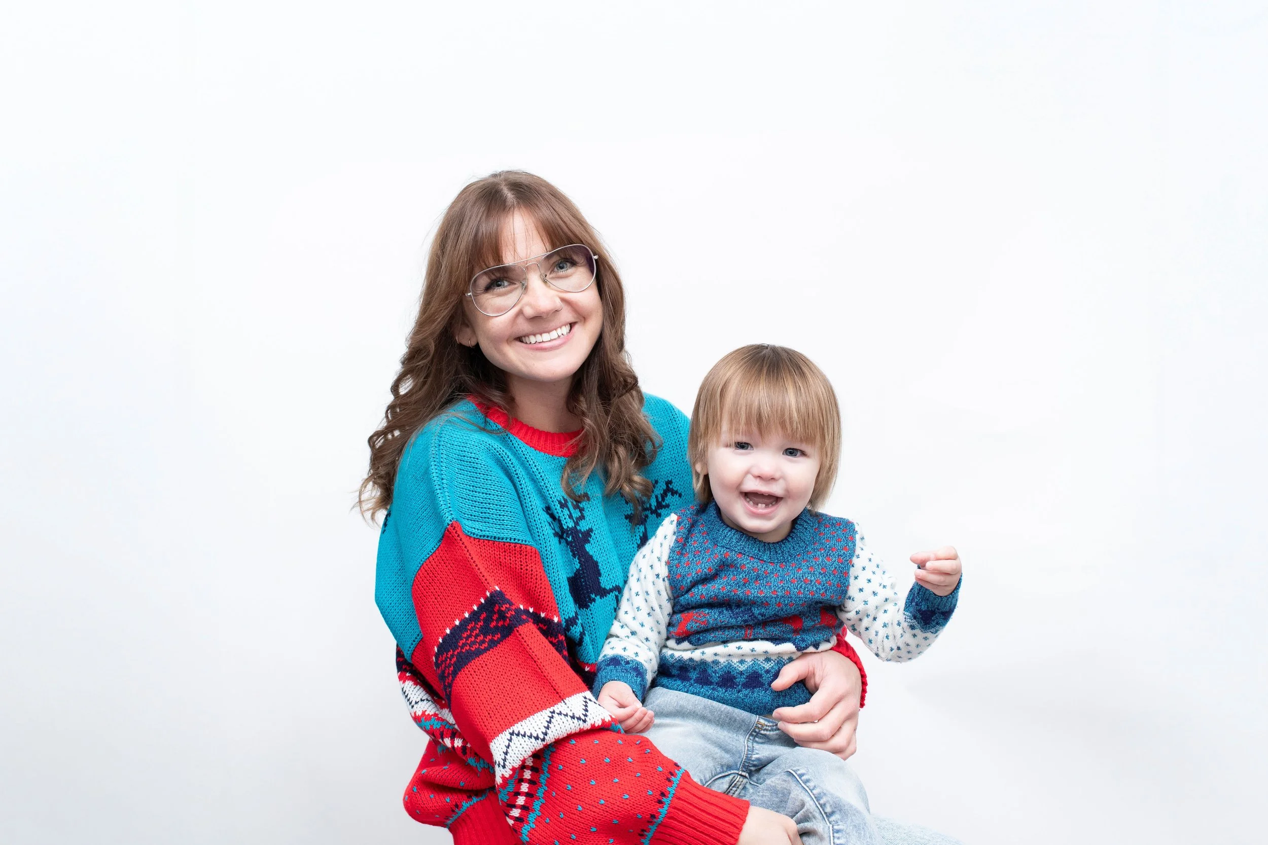 A woman with glasses and a big smile holding a young child on her lap, both wearing Christmas sweaters with festive patterns, sitting against a white background.