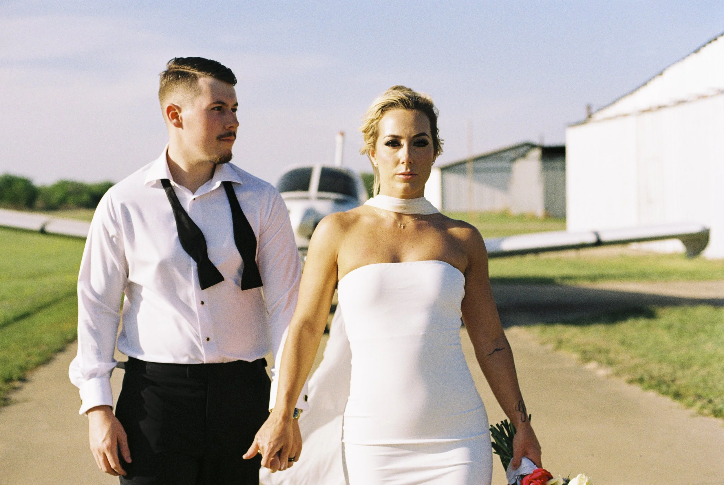 A couple walking outdoors, holding hands. The man wears a white shirt with a loosened black tie, and the woman wears a white strapless dress with a choker neckline. They are walking on a paved path, with a grassy area and buildings in the background.