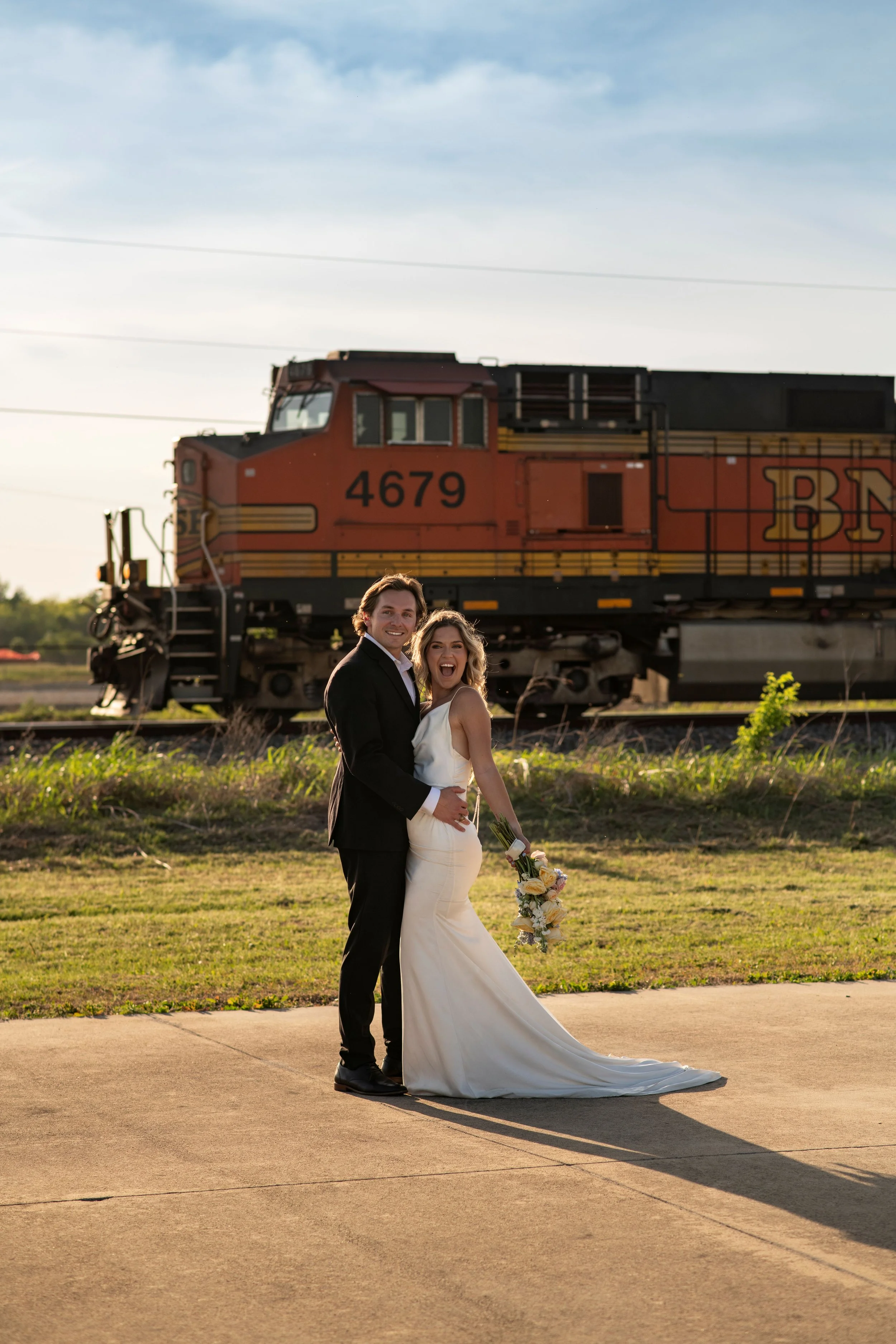 A newlywed couple standing outdoors in front of a moving train on tracks, with a cloudy sky in the background. The bride is wearing a white wedding gown, holding a bouquet, and the groom is in a dark suit.