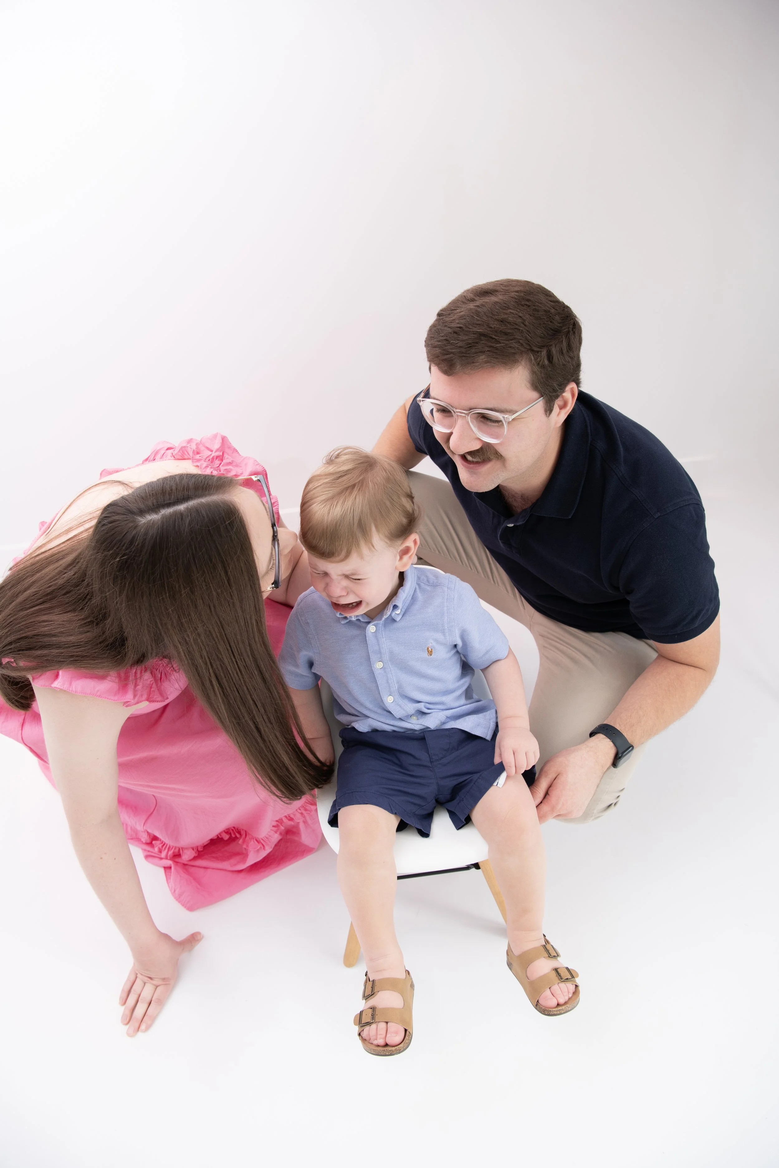 A young boy sitting on a chair crying while a woman and man comfort him, in a studio with a white background.