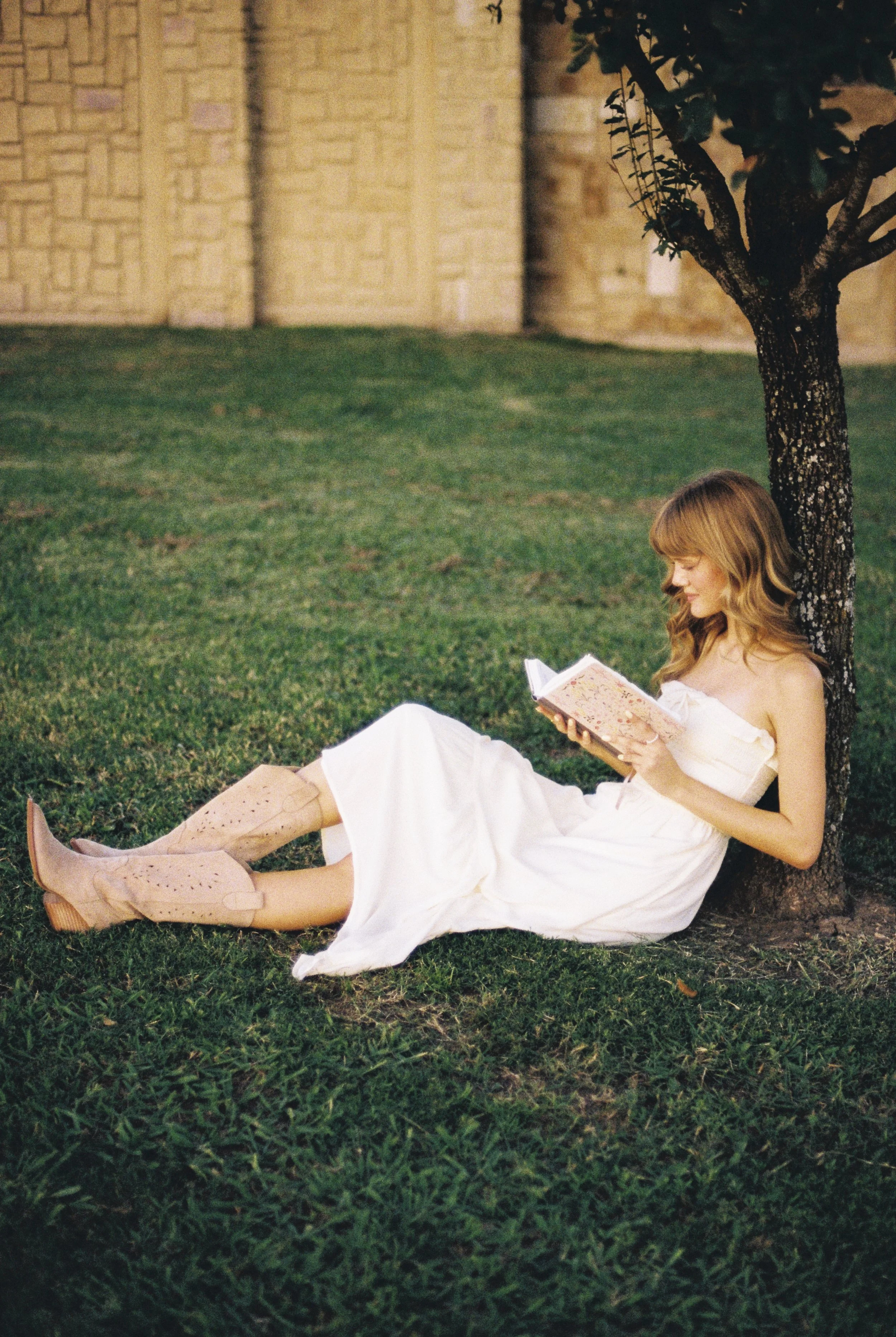 A woman in a white dress and cowboy boots sitting on grass, leaning against a tree while reading a book.