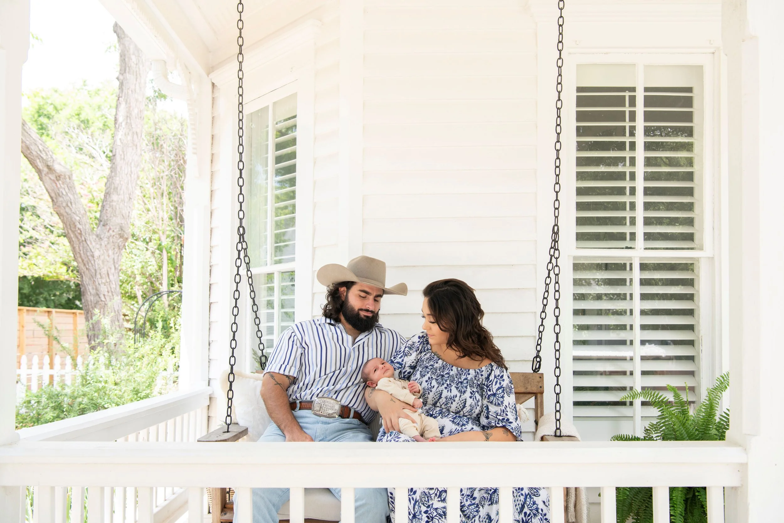 A family sitting on a porch swing, holding a baby, with a white house and green trees in the background.