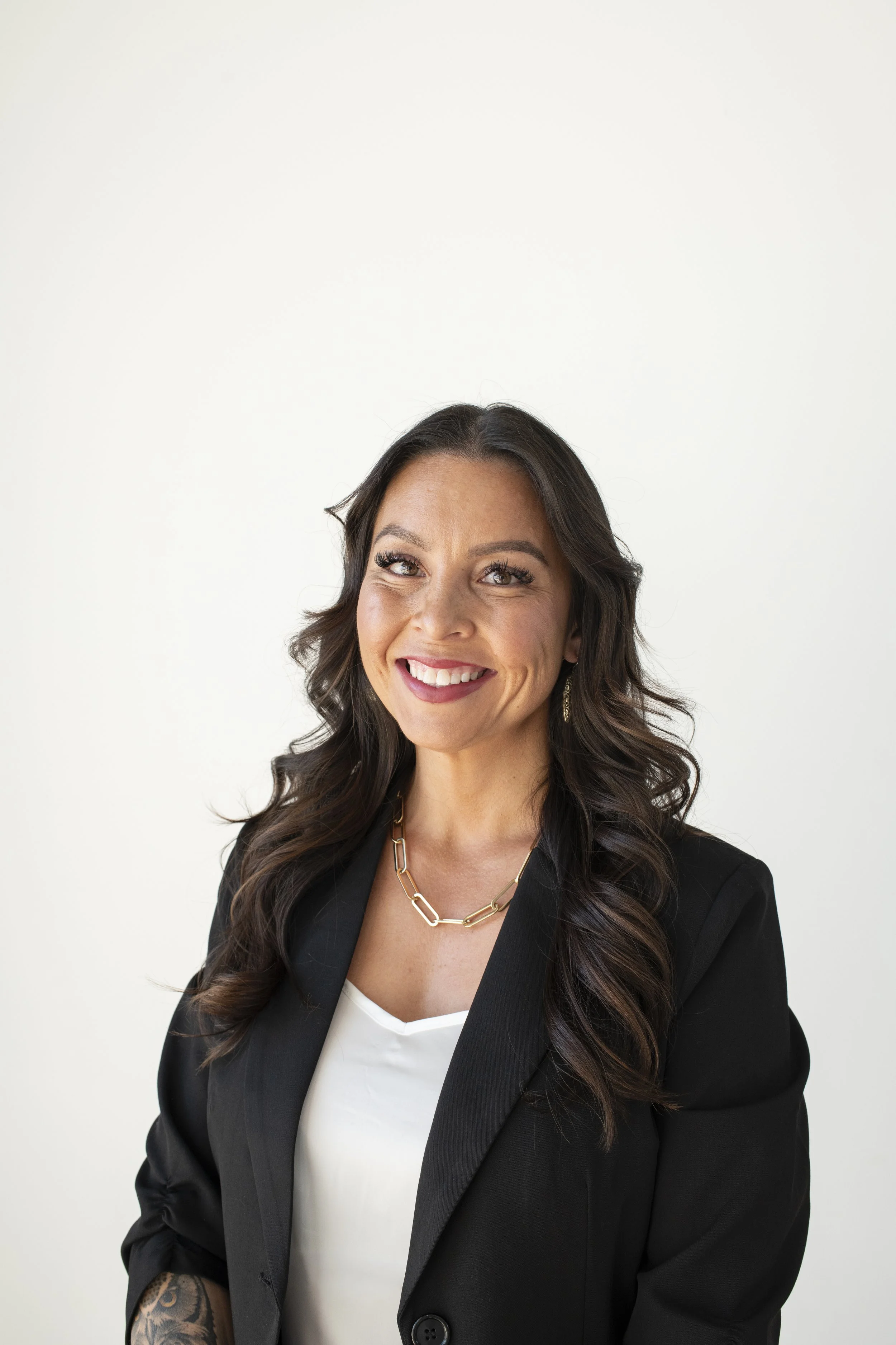 A woman with long dark wavy hair smiling, wearing a black blazer, white top, gold chain necklace, and earrings, standing against a plain white background.