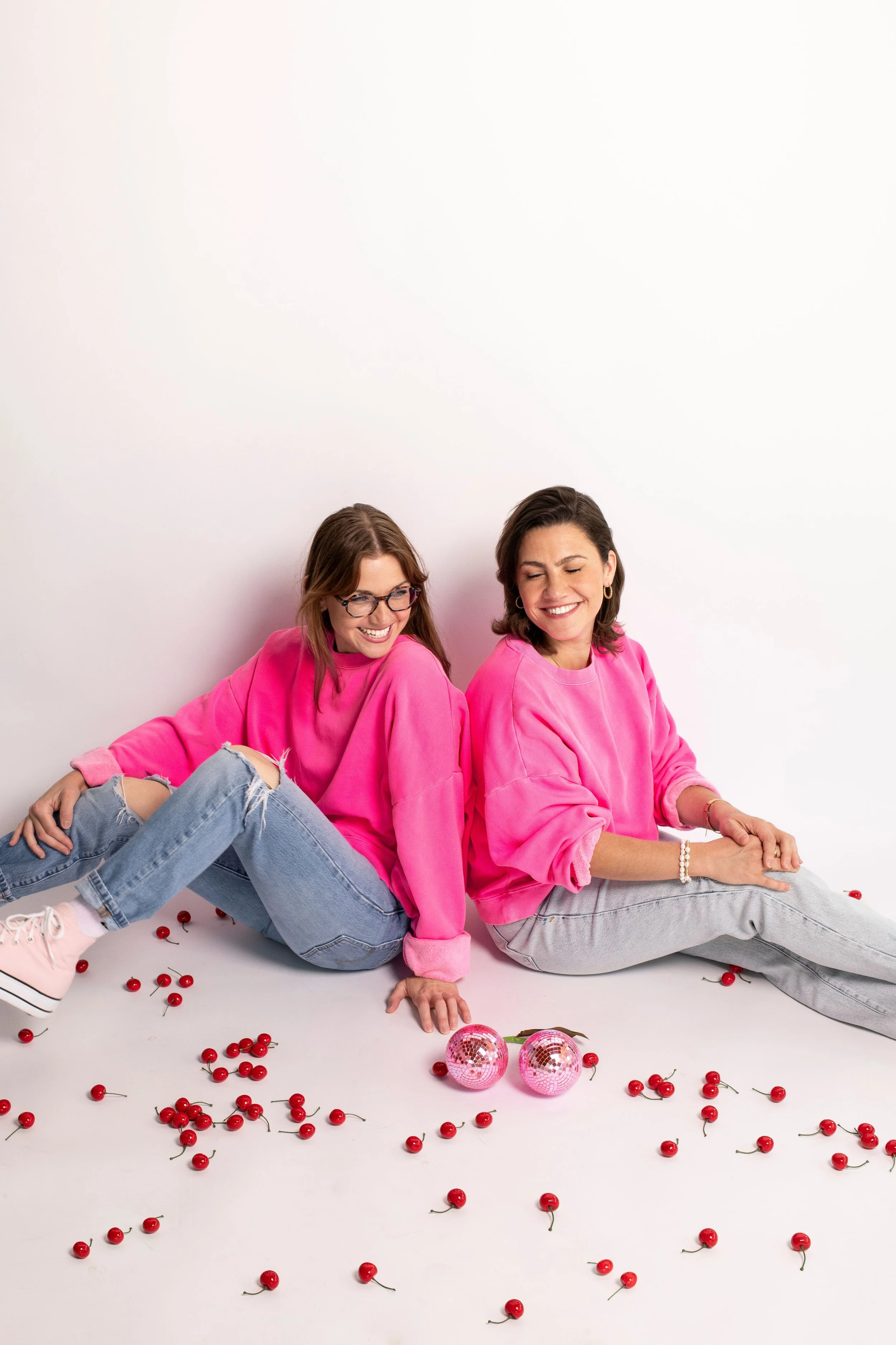 Two women wearing pink sweatshirts and jeans are sitting on the floor surrounded by cherries and two pink disco balls, smiling and laughing together, against a plain white background.