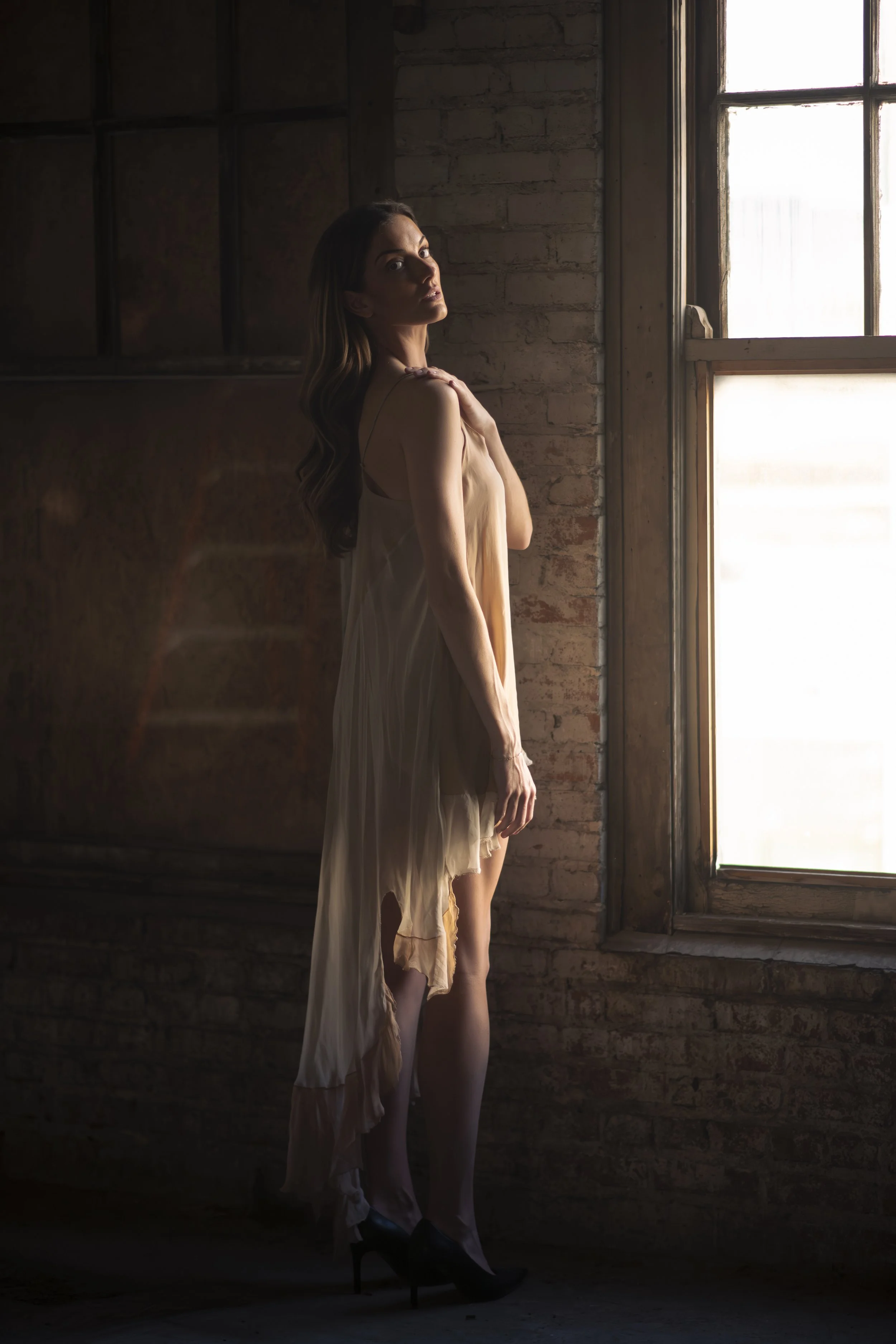 A woman standing near a window with sunlight shining through, wearing a light-colored dress and high heels, in a room with brick walls and wooden elements.