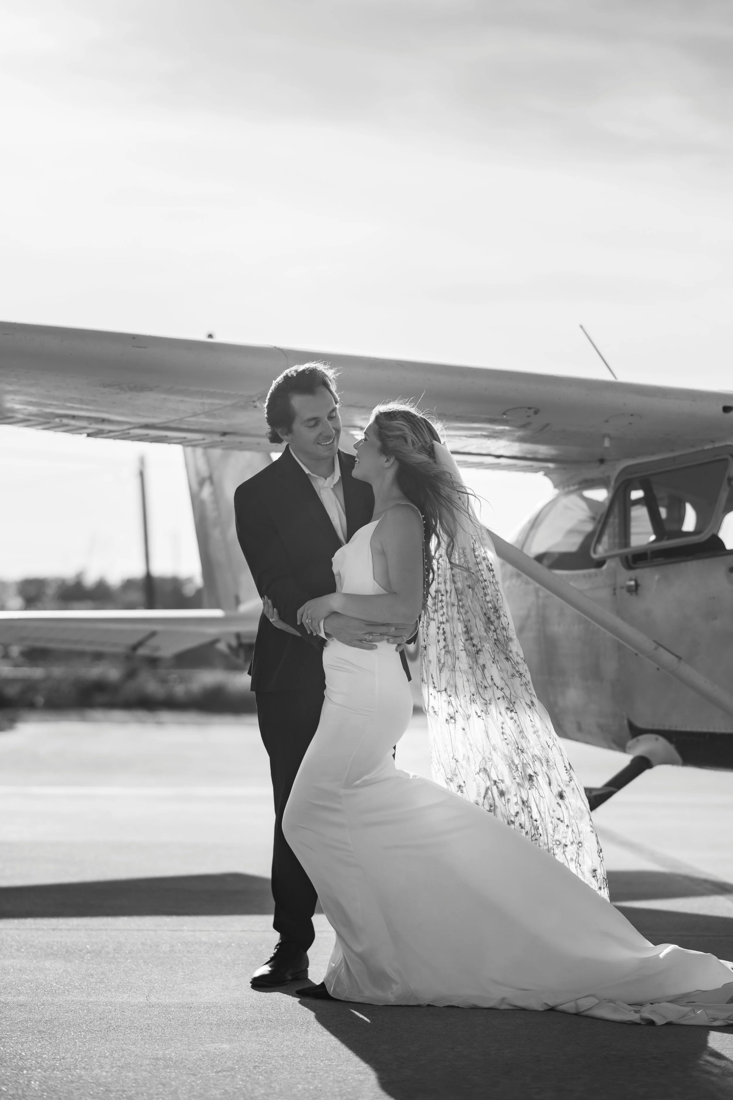 A couple, dressed in wedding attire, sharing a dance or intimate moment near a small airplane on a tarmac.