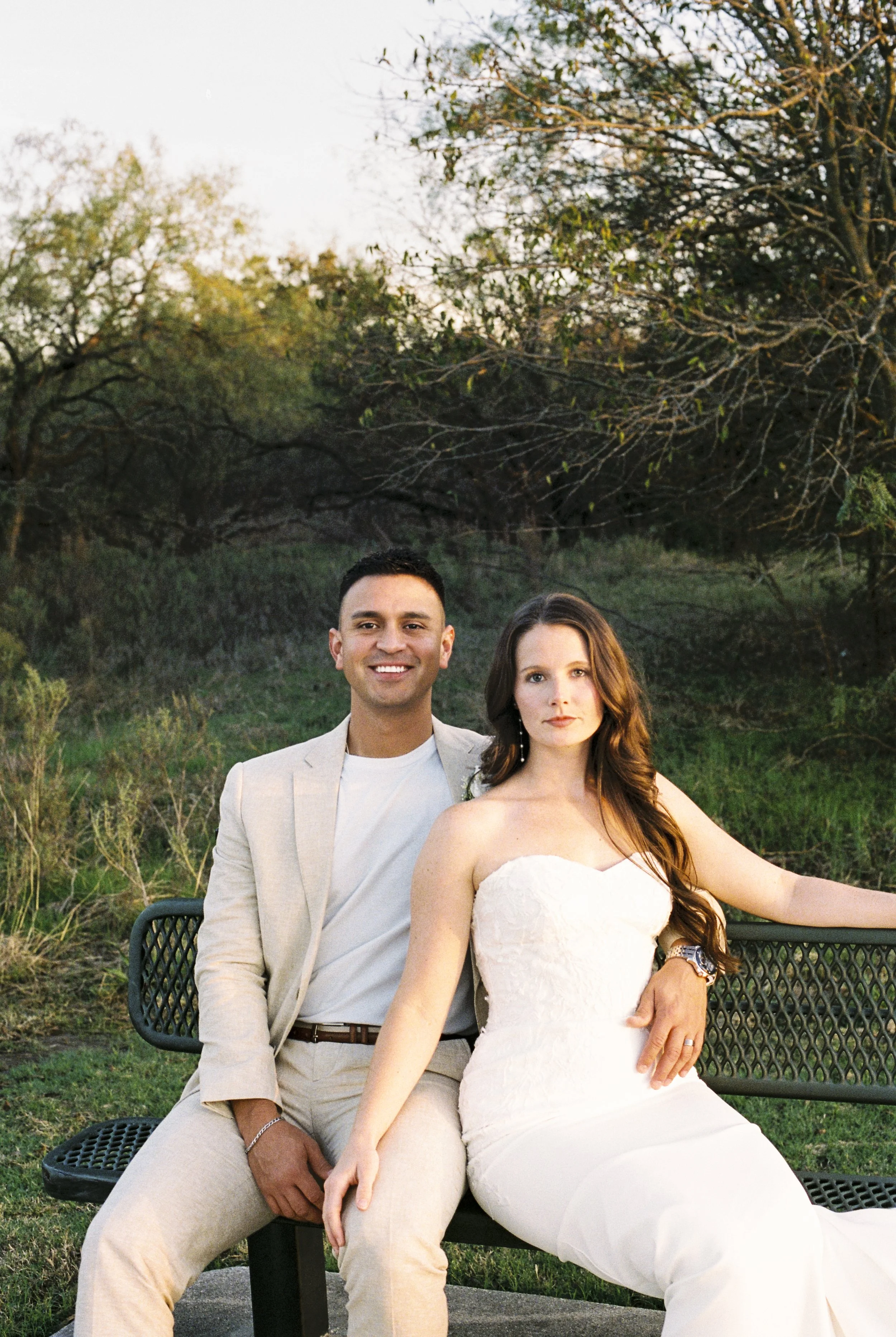A smiling man and a serious woman sit on a park bench outdoors during sunset. The man wears a light-colored suit with a white shirt, and the woman wears a white strapless wedding dress. They are surrounded by trees and greenery.
