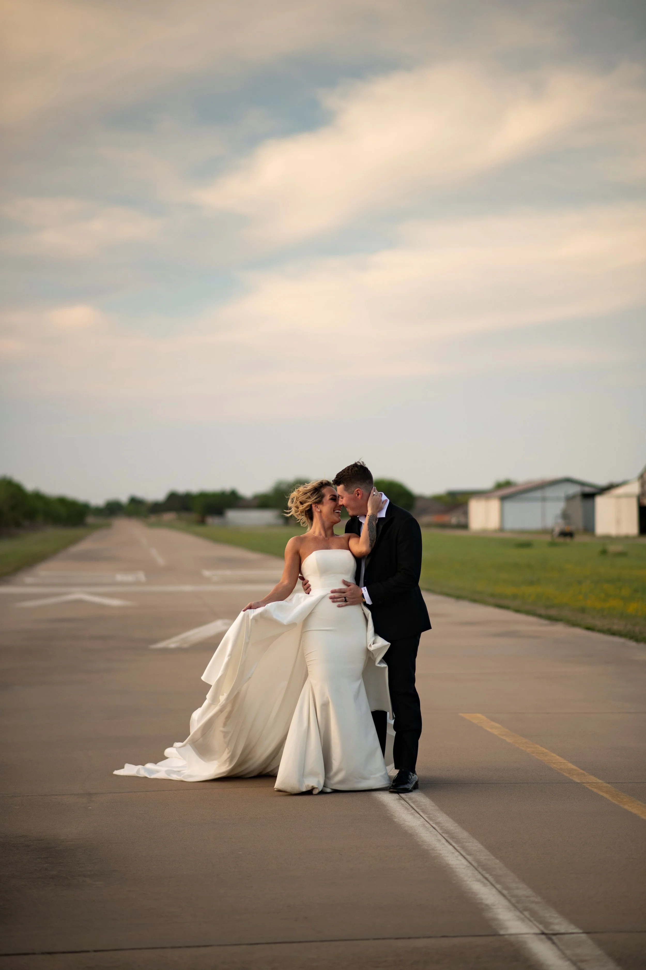A bride and groom standing in the middle of an empty runway or road, sharing a tender moment with their foreheads touching, outdoors with a cloudy sky overhead.
