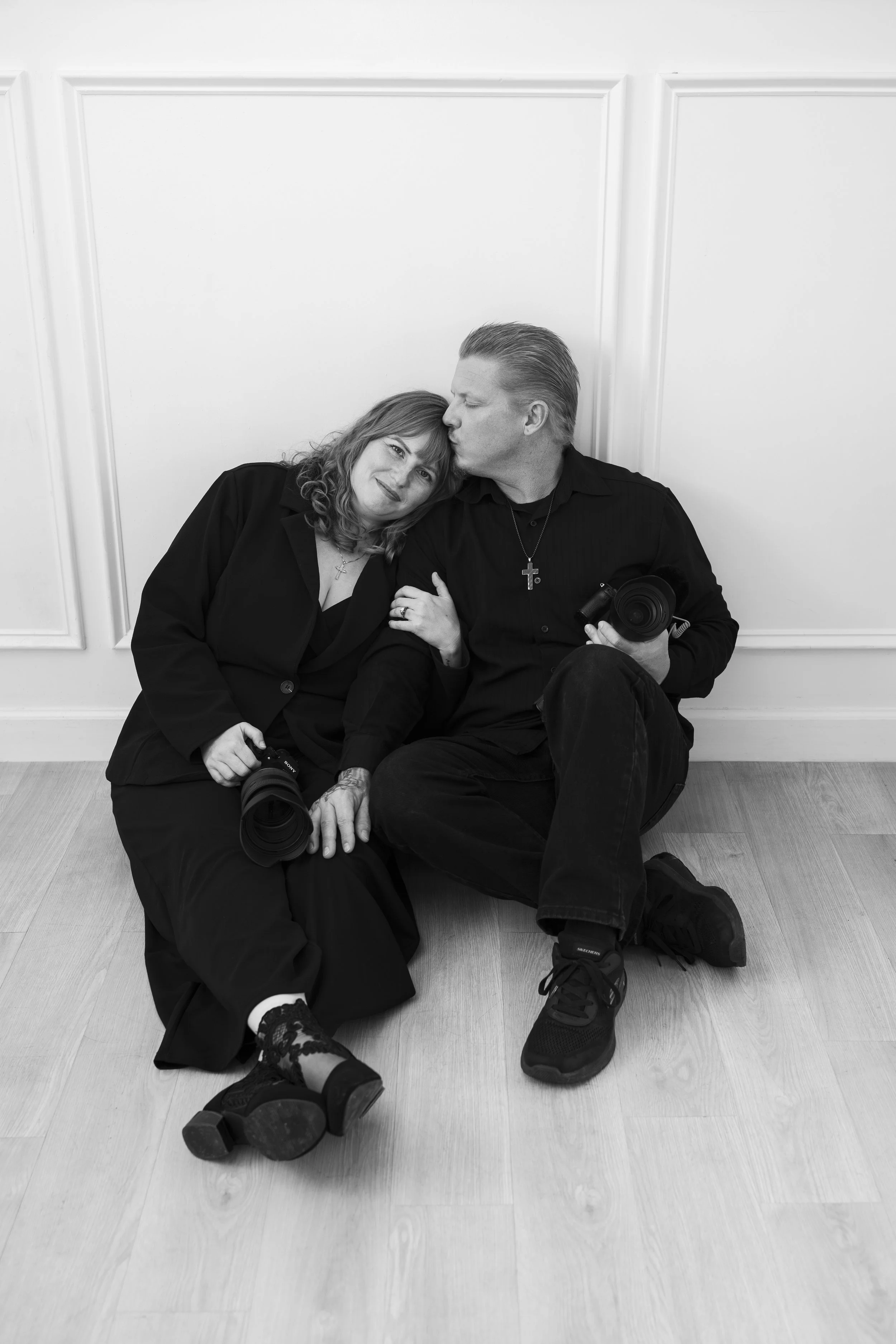 A black and white photo of a man and woman sitting on the floor against a white wall with decorative paneling. The woman is leaning her head on the man's shoulder, both holding cameras with friendly expressions. The woman has curly hair, and the man 