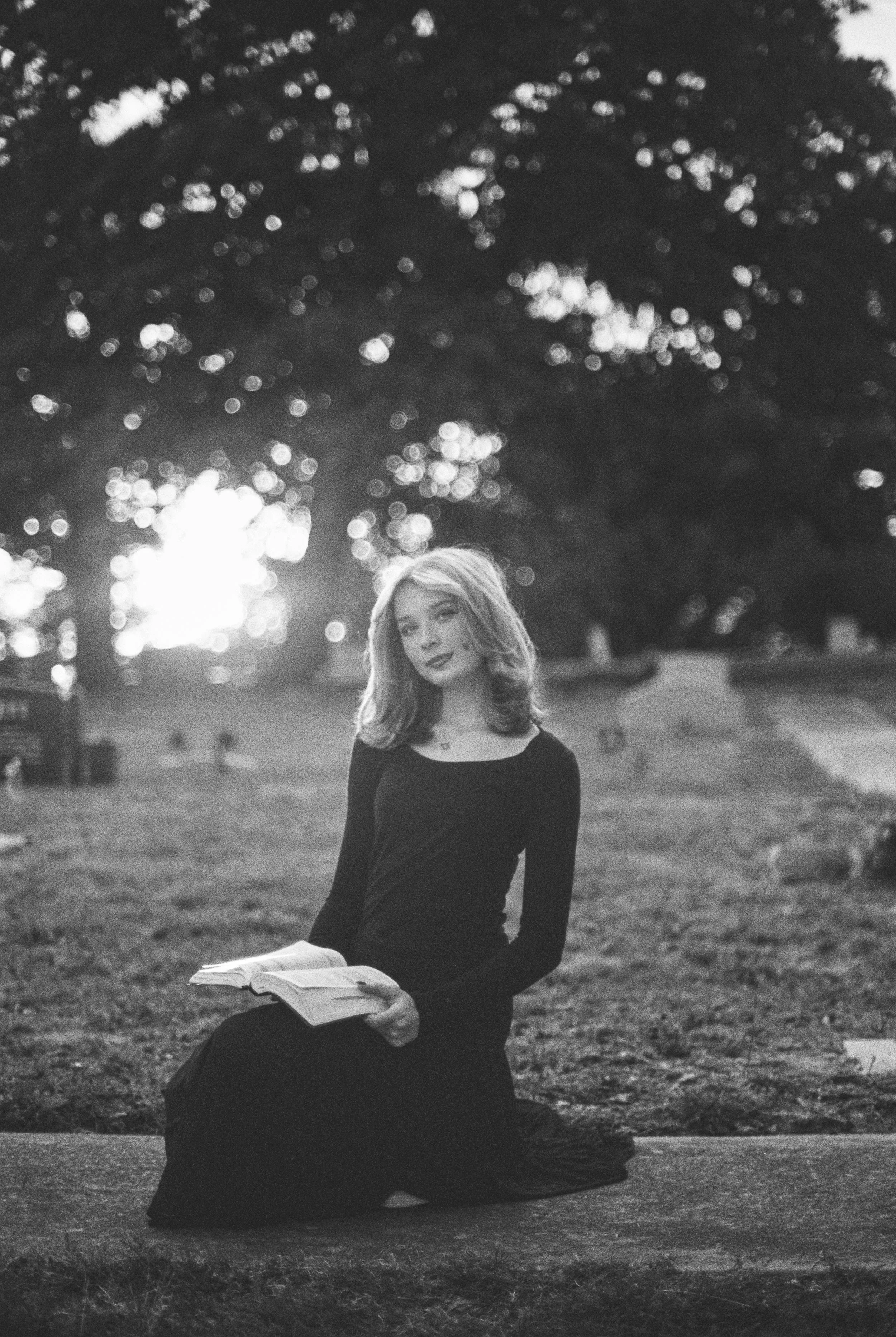 A young woman with shoulder-length hair, wearing a long black dress, sits on a curb in a park at sunset, holding an open book in her lap, with a large tree in the background.