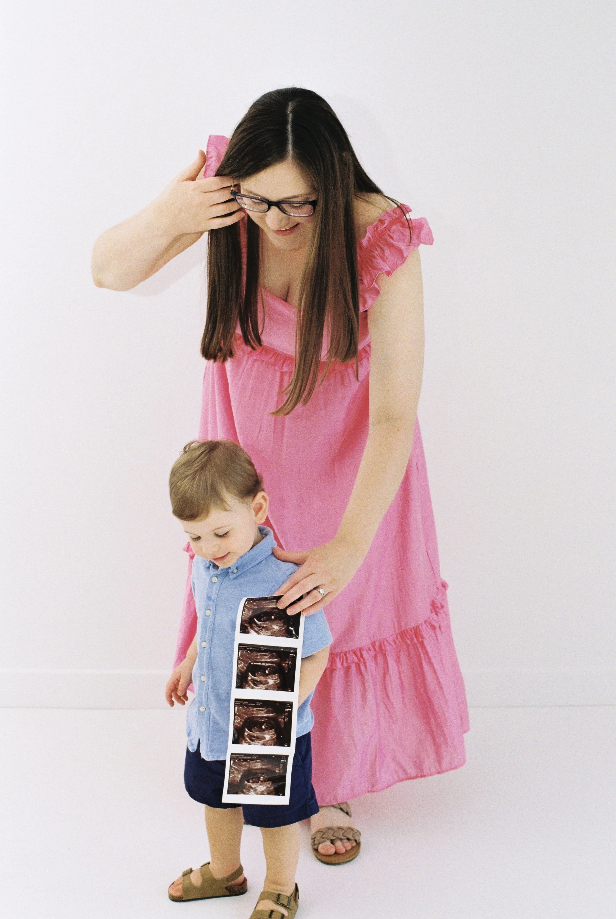 A woman in a pink dress with glasses is smiling and touching her head, standing next to a young boy holding ultrasound images. The boy is wearing a blue shirt, navy shorts, and sandals, looking at the ultrasound images.