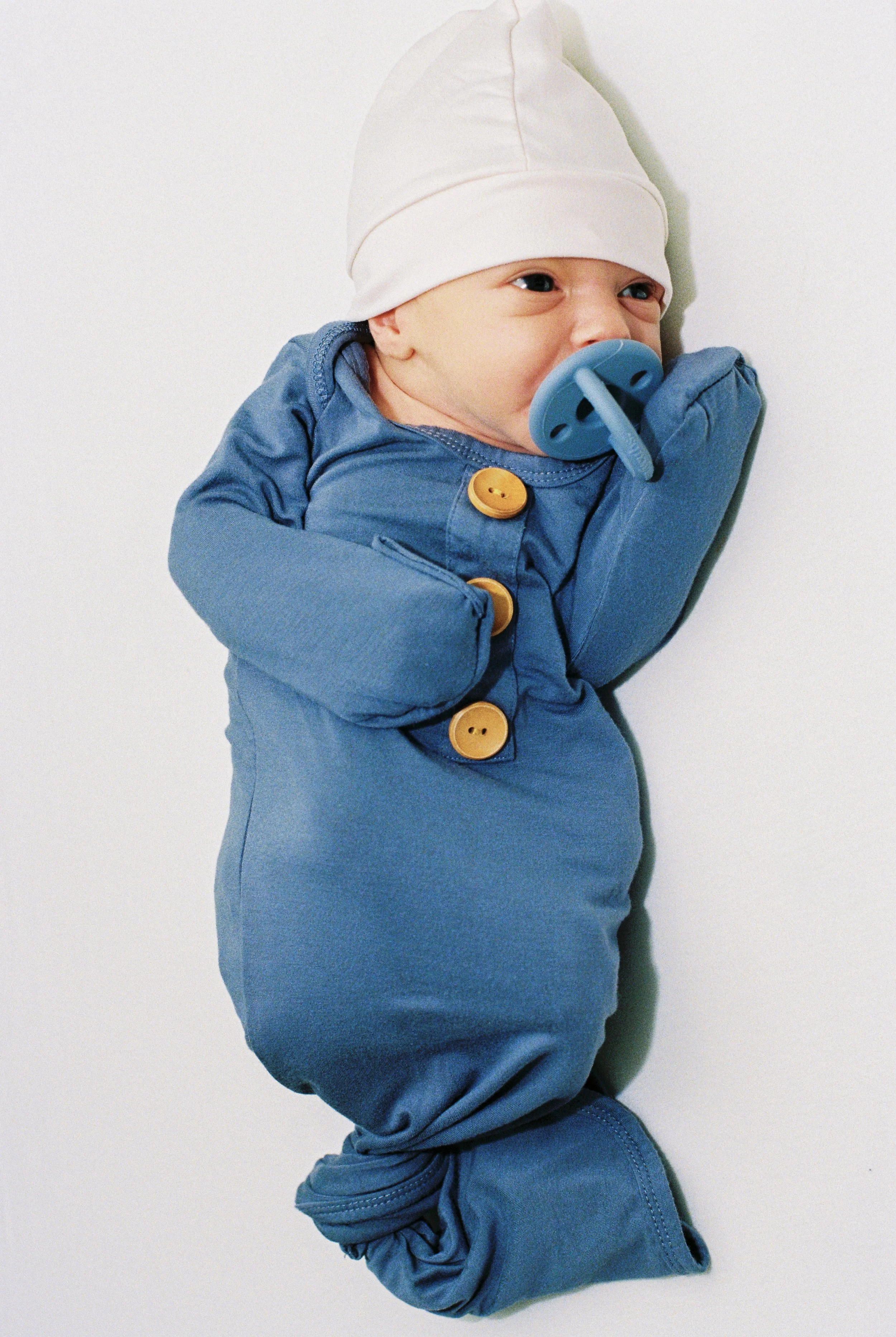 A baby lying on a white surface, wearing a blue onesie, a white hat, and a pacifier in their mouth.