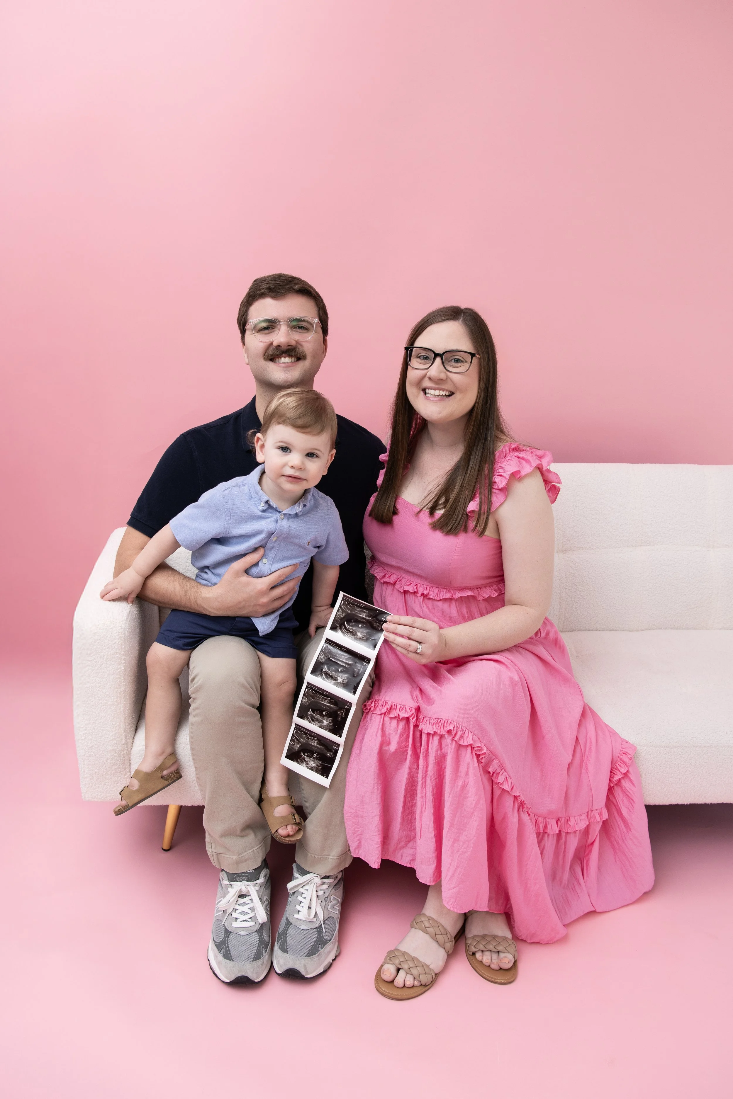 A family of three sitting on a white sofa against a pink background, holding ultrasound pictures, with the mother in a pink dress, the father in a dark shirt, and a toddler boy in a blue shirt and beige shorts sitting on his father's lap.