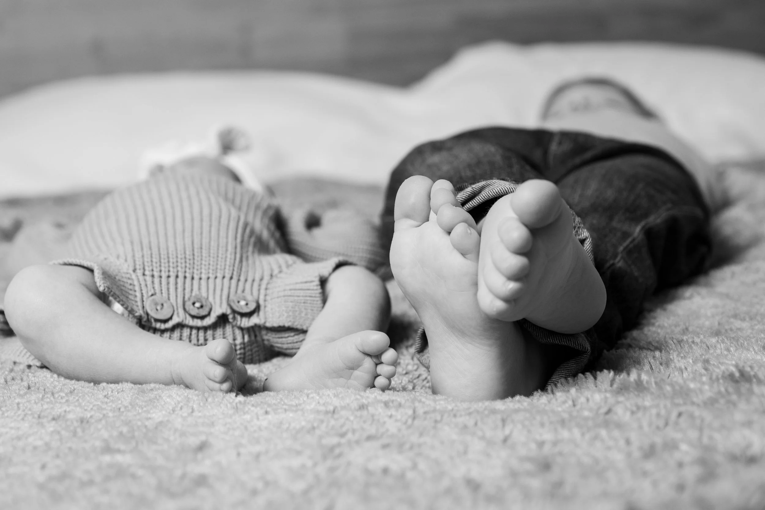 Black and white photo of two children lying on a soft surface, with one child's feet in the foreground and another child's face and body partially visible in the background.
