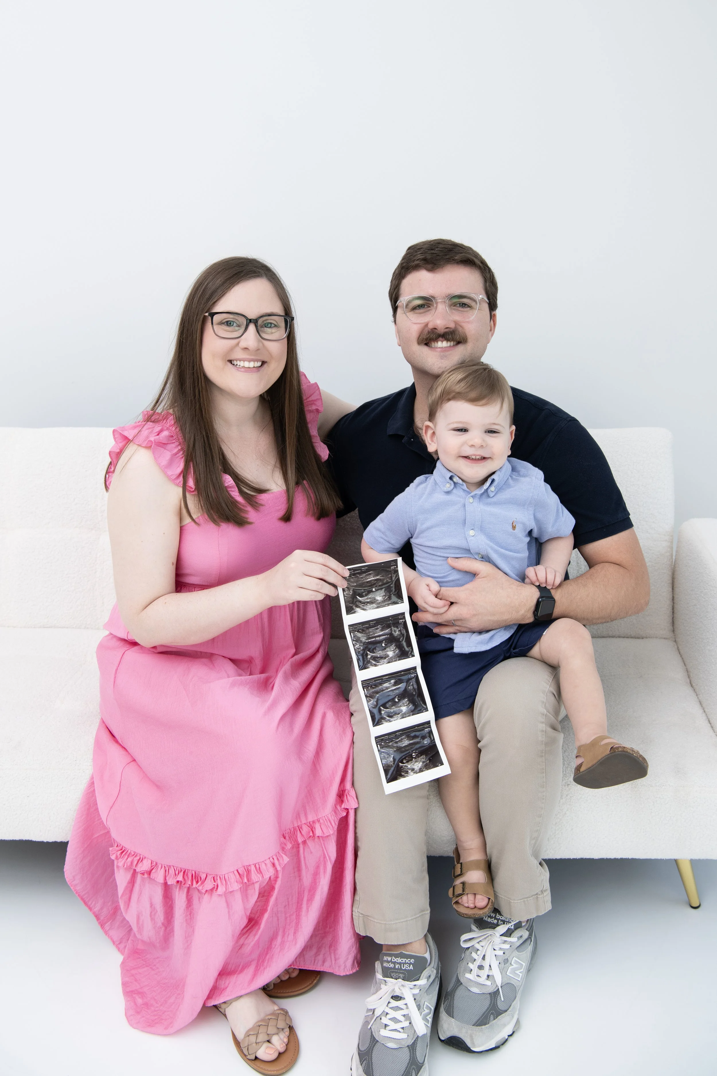 A couple with their young son sitting on a white couch, holding ultrasound images of a pregnancy, celebrating expecting a new baby.