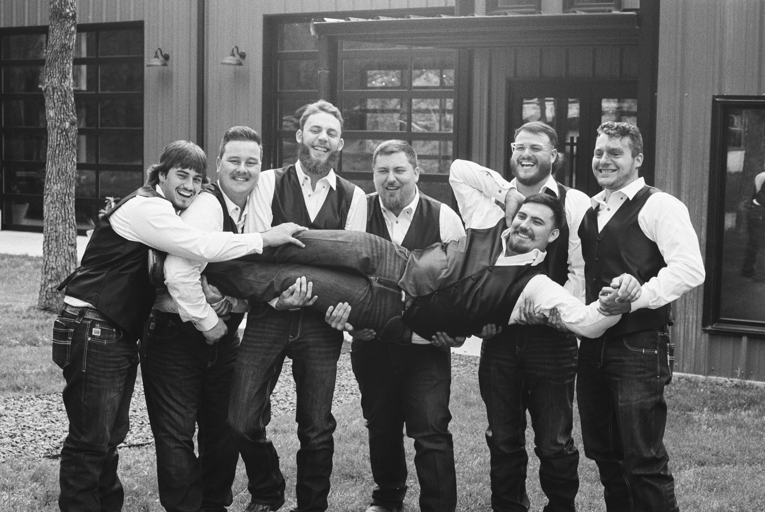 A group of grooms men wearing dress shirts and vests, holding a man horizontally in their arms, all smiling and laughing outside in front of a modern building with large windows.
