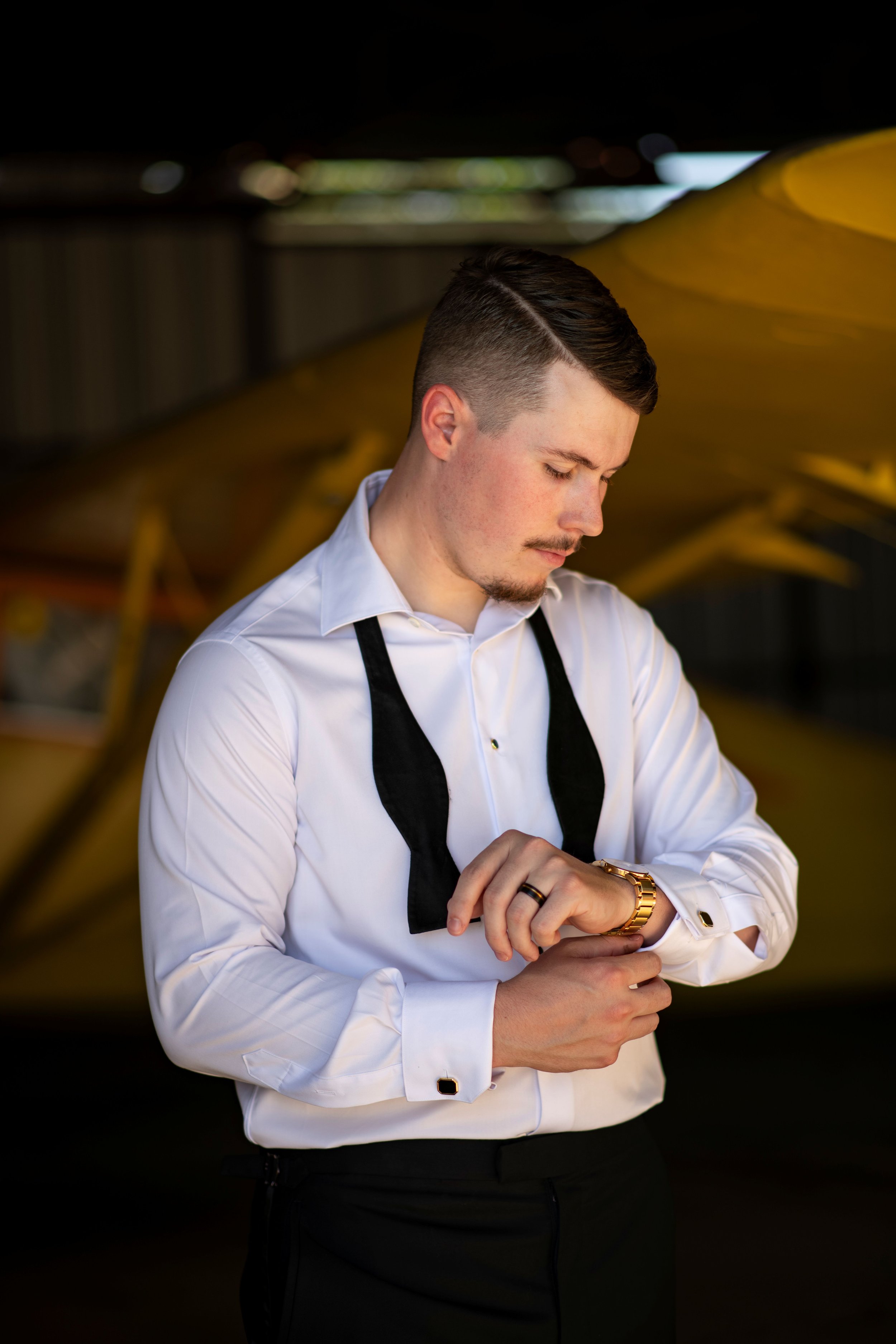 Young man in a tuxedo with a white shirt and black bow tie, looking at his watch in a dimly lit area with yellow and black objects in the background.