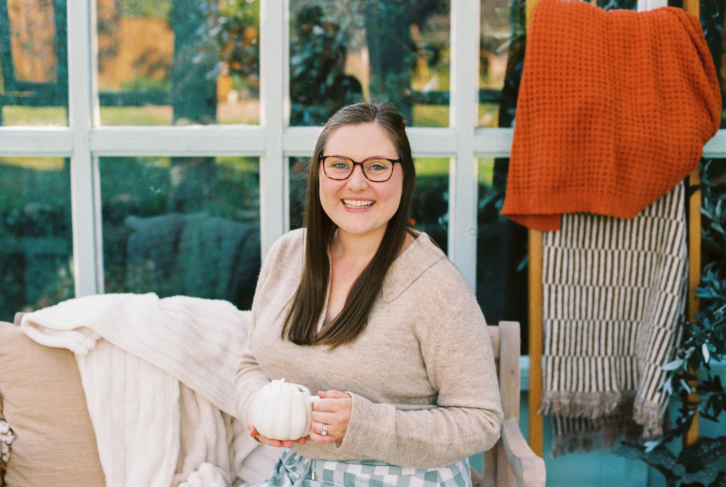 A woman with glasses smiling, sitting indoors near a window, holding a small white pumpkin, with a cozy blanket and a cushion nearby.