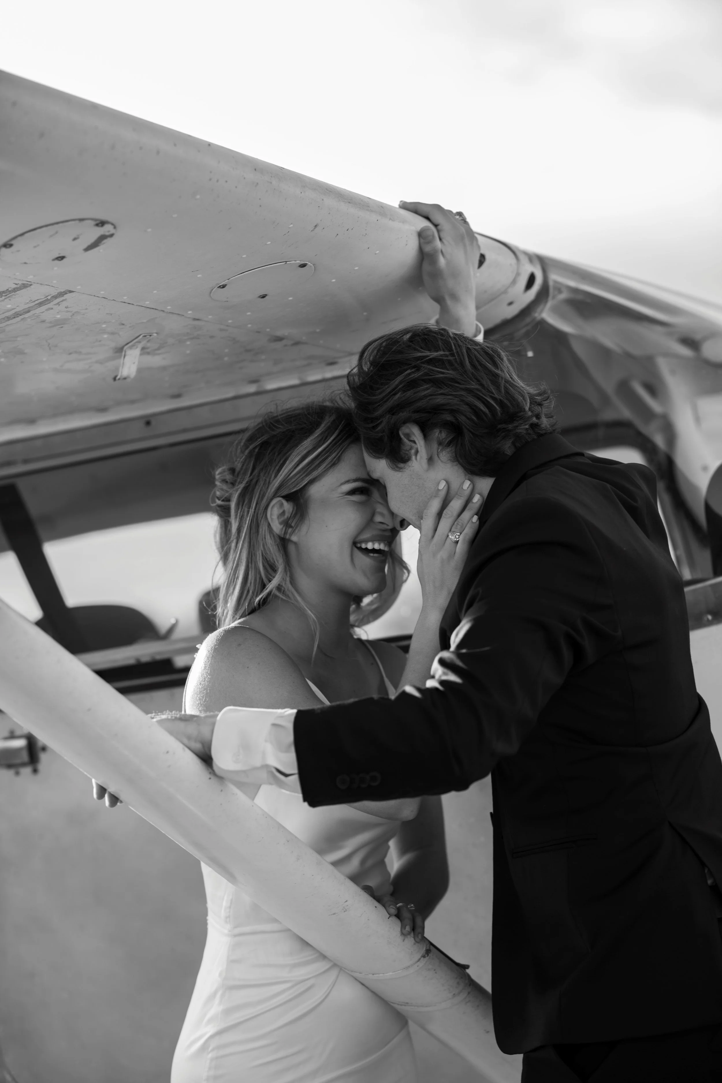A couple dressed in wedding attire sharing a kiss under an airplane wing on a cloudy day.
