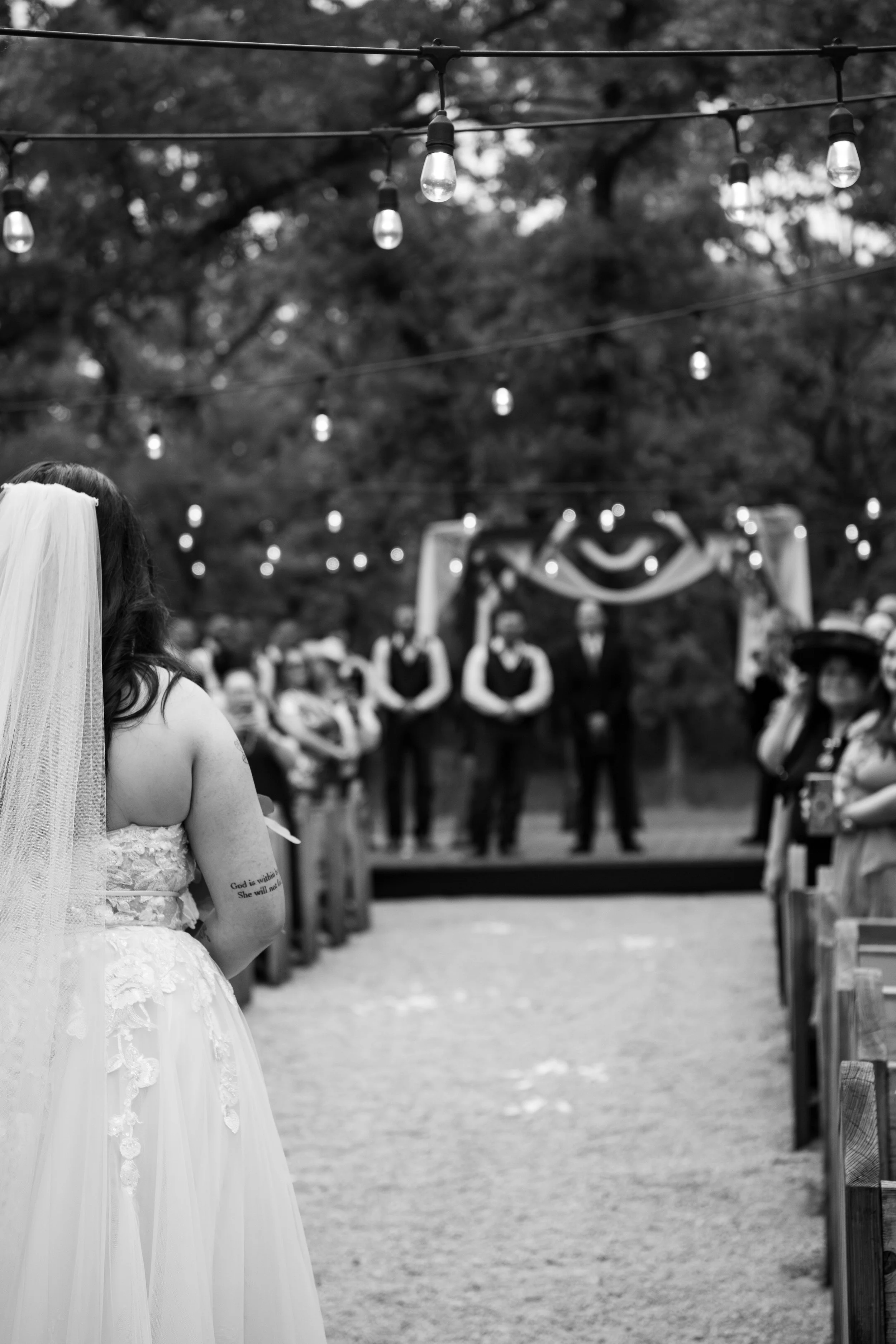 Black and white image of a bride in a wedding dress and veil standing outdoors, facing away, with wedding guests and string lights in the background at a wedding ceremony.