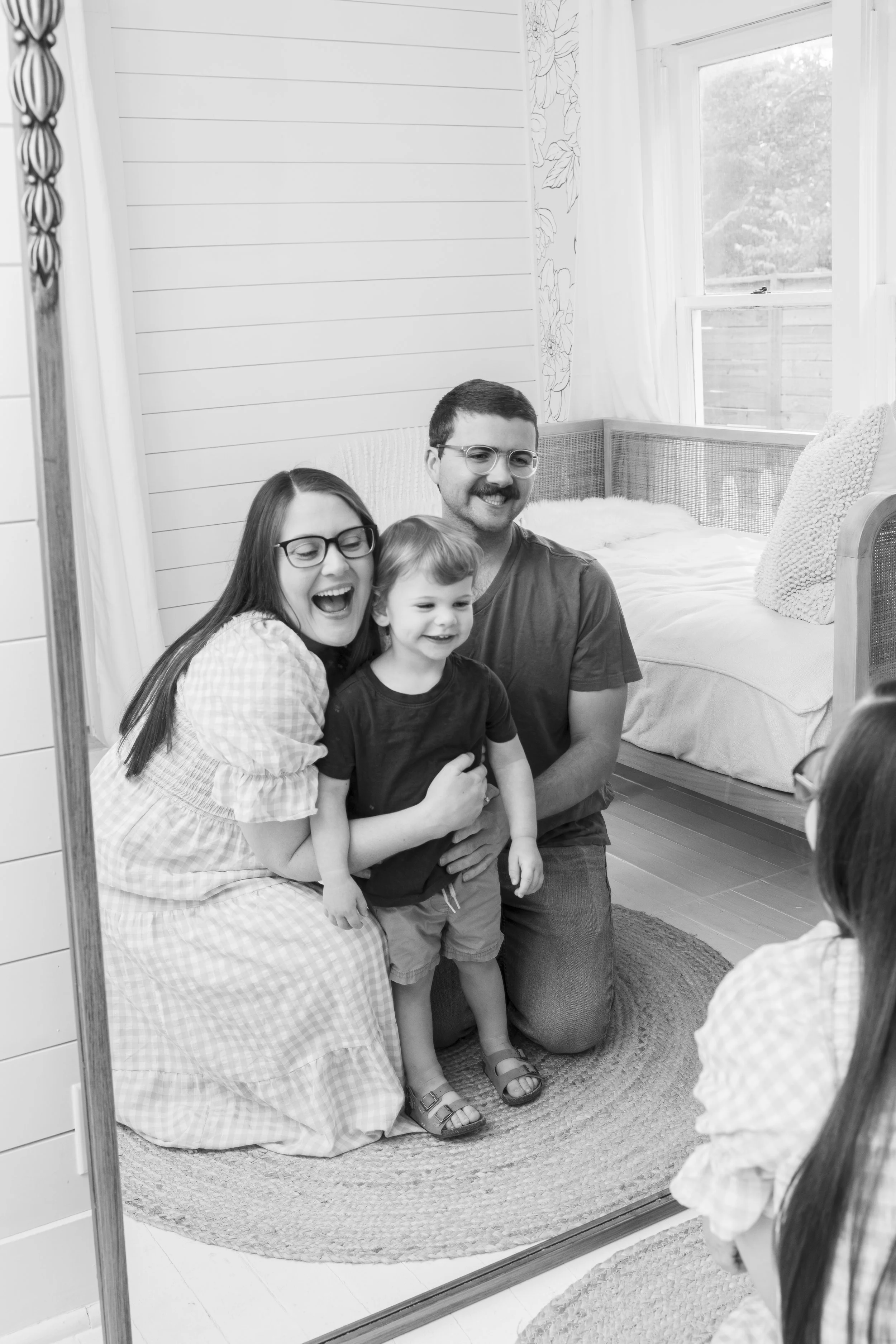 A family of four taking a selfie inside a bedroom with a large mirror. The family includes a woman with long dark hair and glasses, a man with glasses, a young boy, and a woman with glasses. The background shows a bed, window, and some curtains.