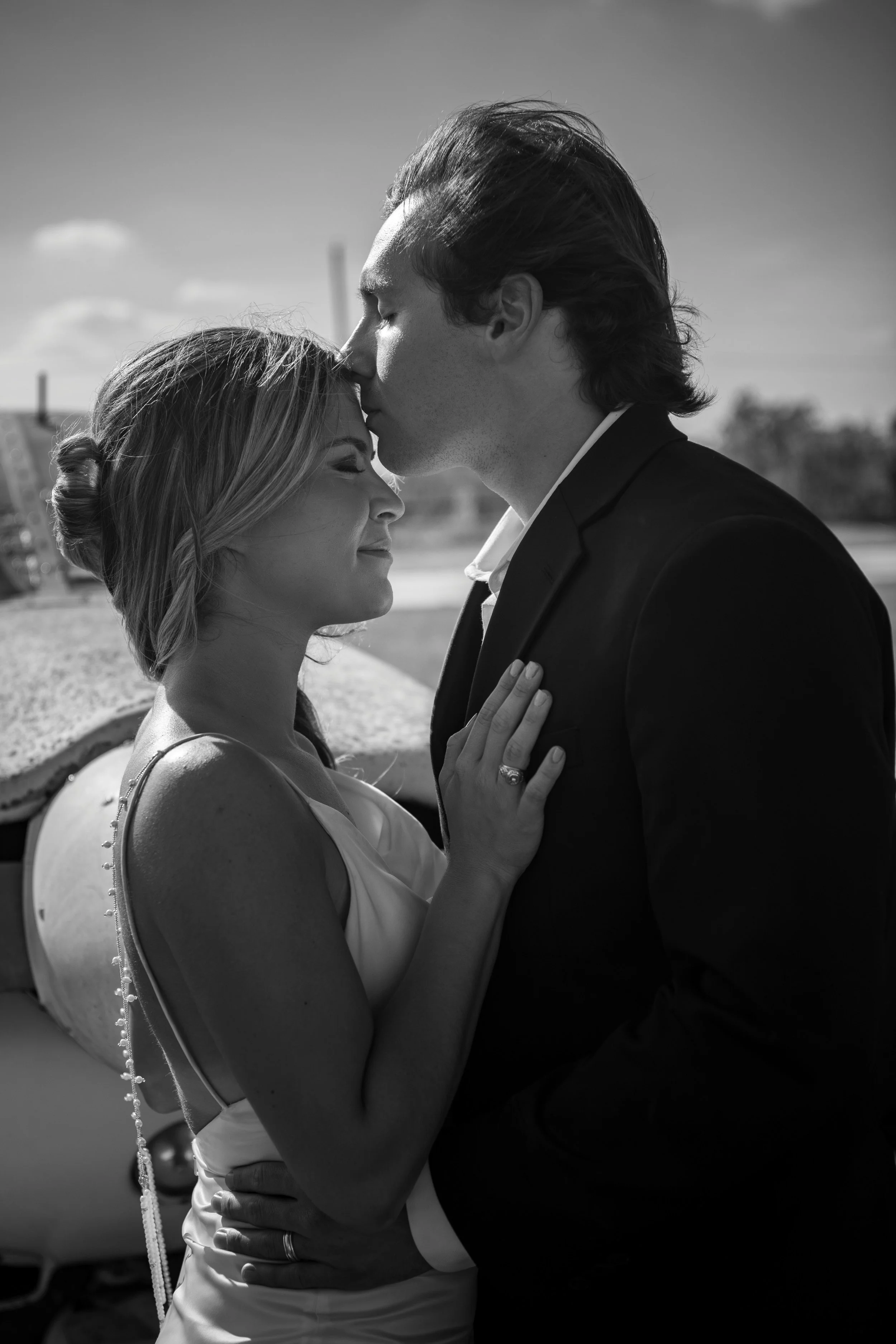 A black and white photo of a couple embracing outdoors, with the man kissing the woman's forehead and her eyes closed, smiling softly.