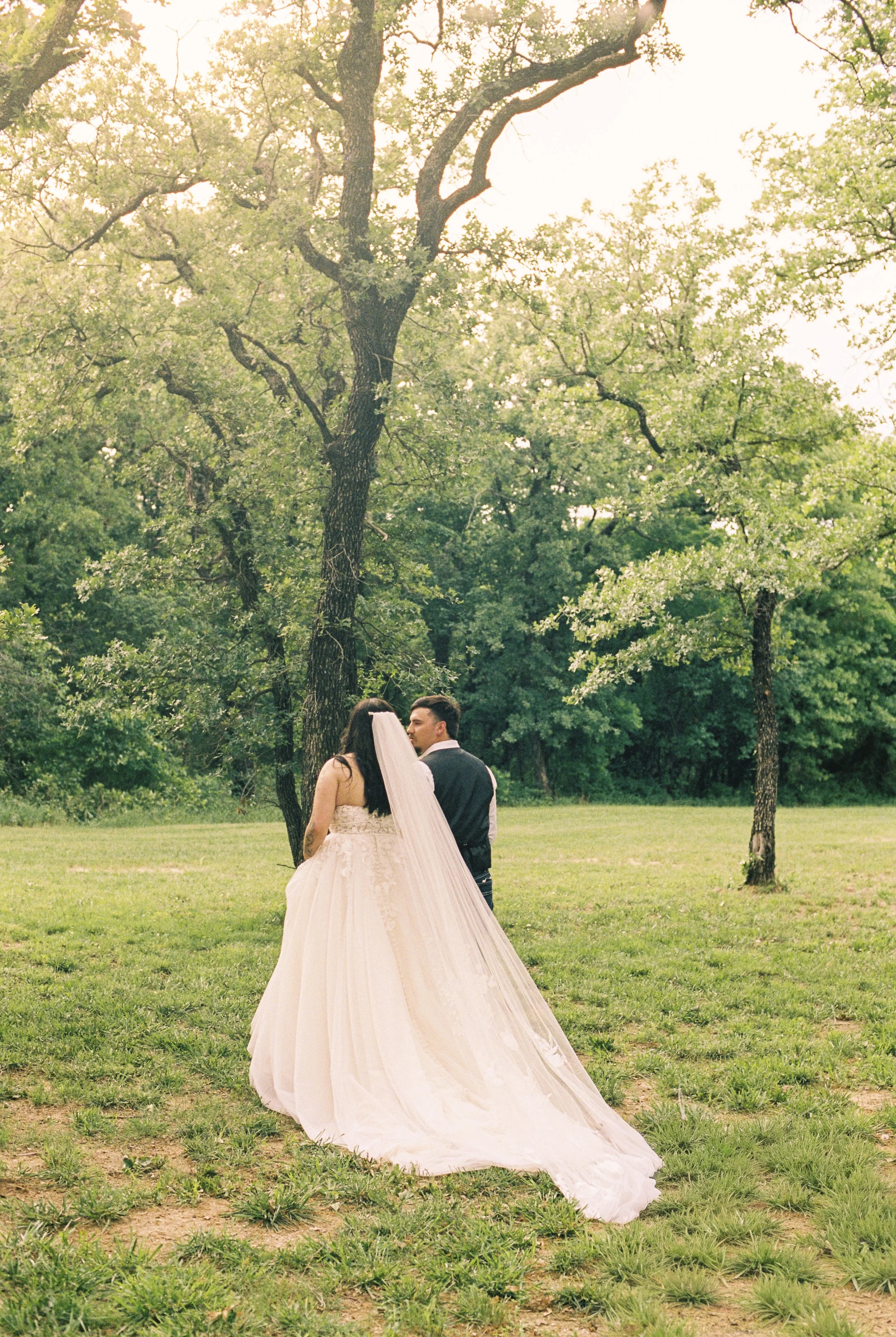A bride and groom standing outdoors on a grassy field, surrounded by trees, during their wedding ceremony. The bride wears a white wedding dress with a long train and veil, and the groom wears a dark suit. They are facing each other, sharing a moment