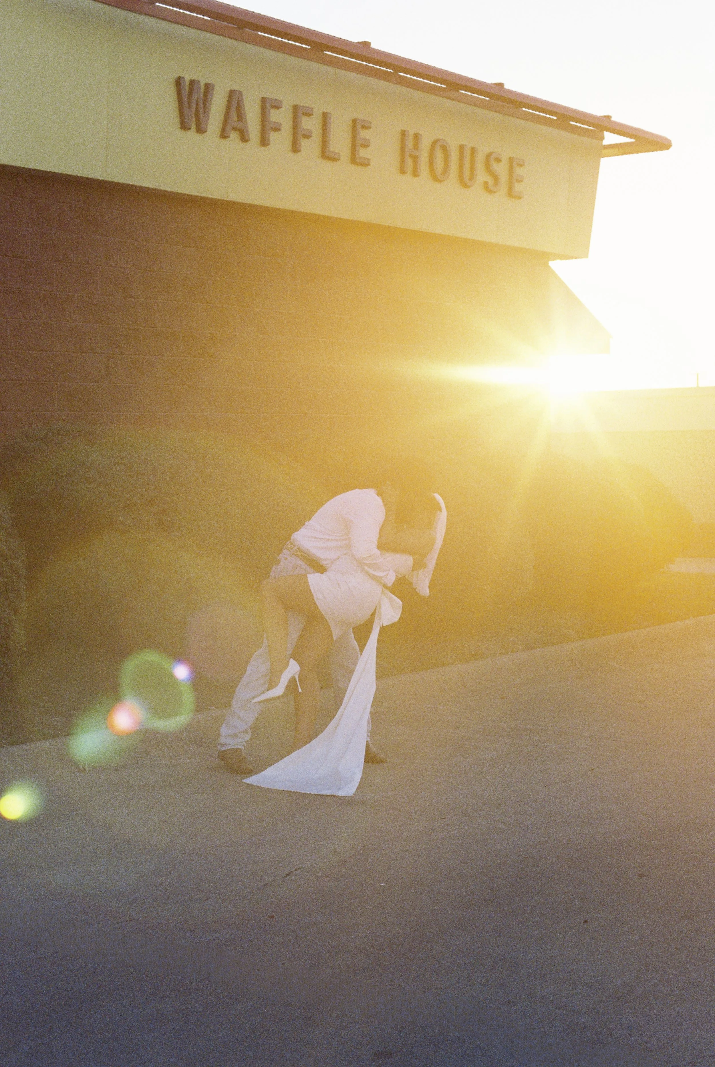 A couple in wedding attire kiss beneath the sign of a Waffle House, with sunlight creating a lens flare.