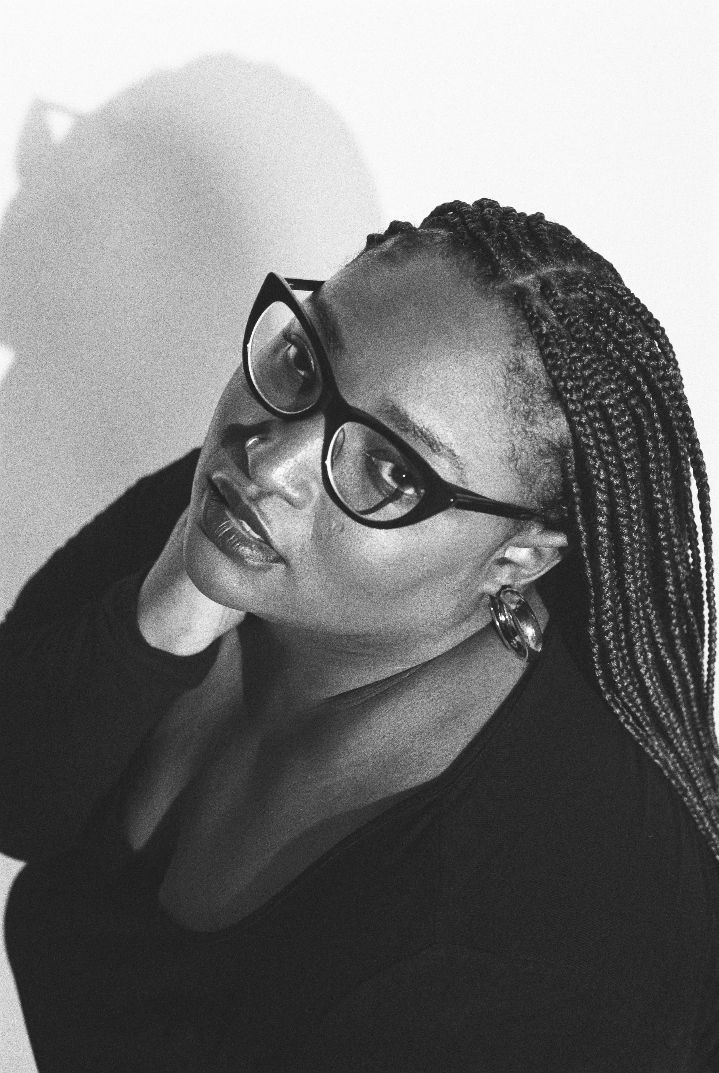 Black and white portrait of a woman with braided hair and glasses, looking at the camera with her hand resting on her neck.