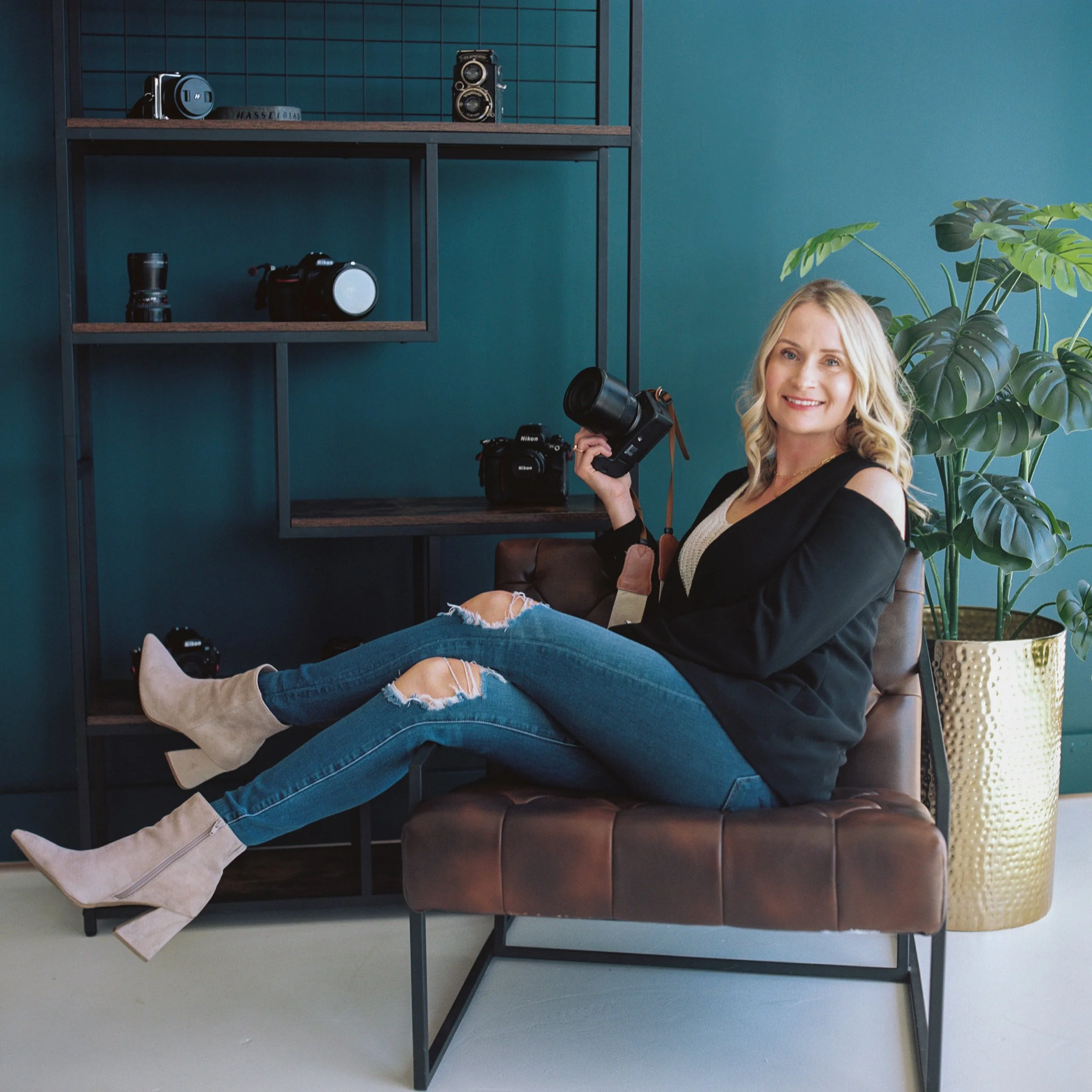 A woman with blonde hair sitting on a brown leather bench, holding a camera, wearing ripped jeans and beige boots, in a room with dark teal walls and a large potted plant, with camera gear on shelves behind her.