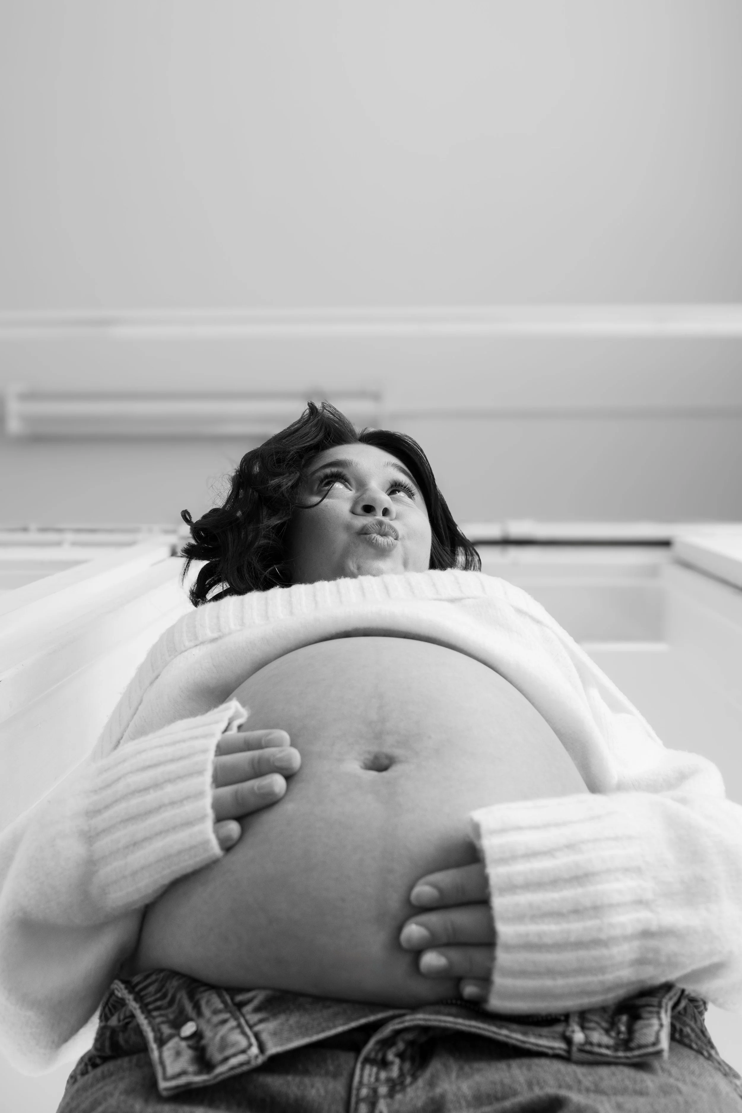 Black and white photo of a pregnant woman looking up, with her hands on her belly, wearing a sweater, standing indoors.